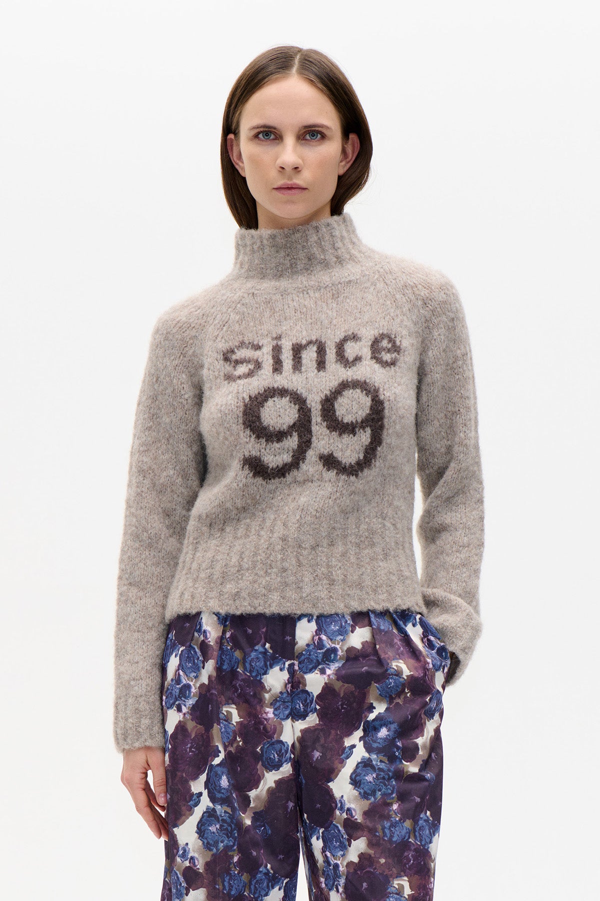 Model wearing a fuzzy taupe high-neck sweater with “since 99” lettering and purple-blue floral trousers, front view.