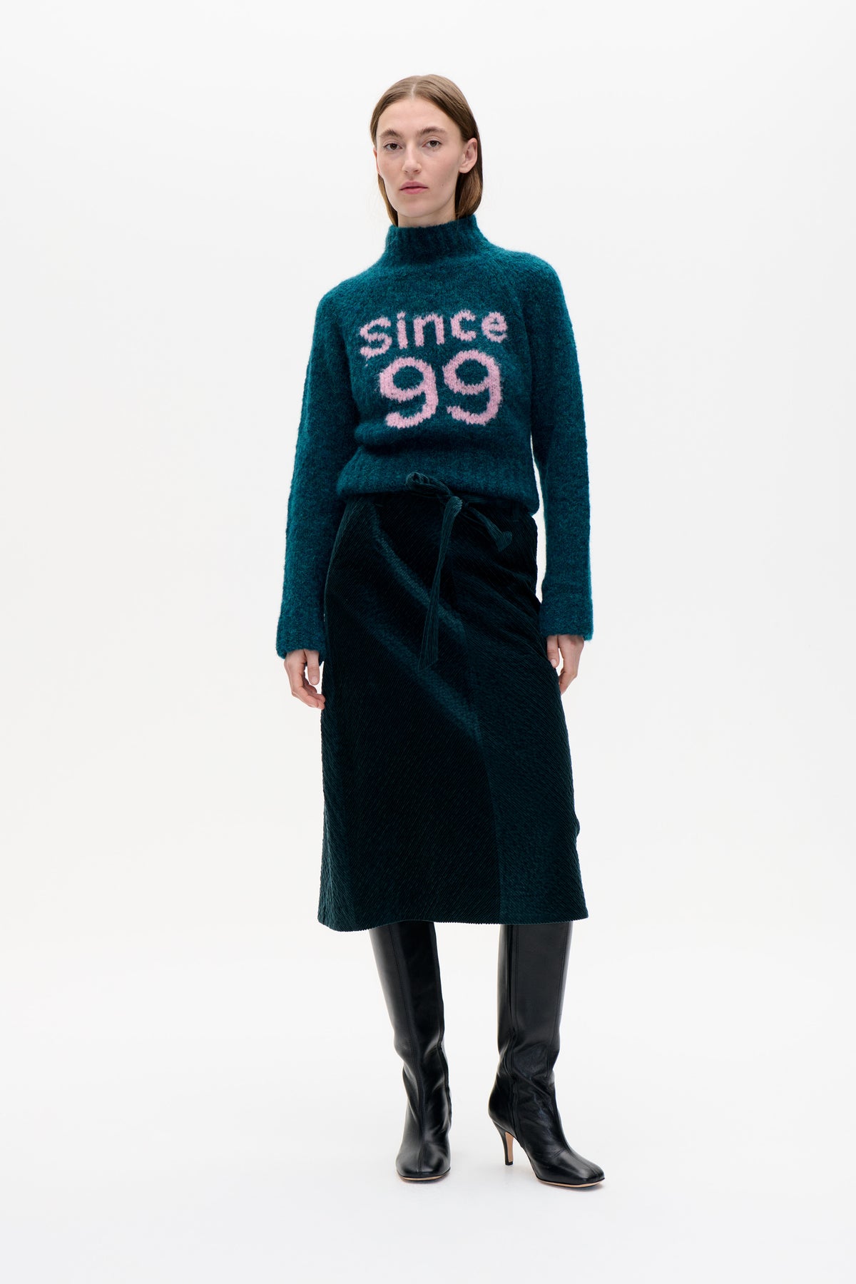 Model wearing a teal high-neck sweater with pink “since 99” lettering, a dark corduroy midi skirt and black knee-high boots.