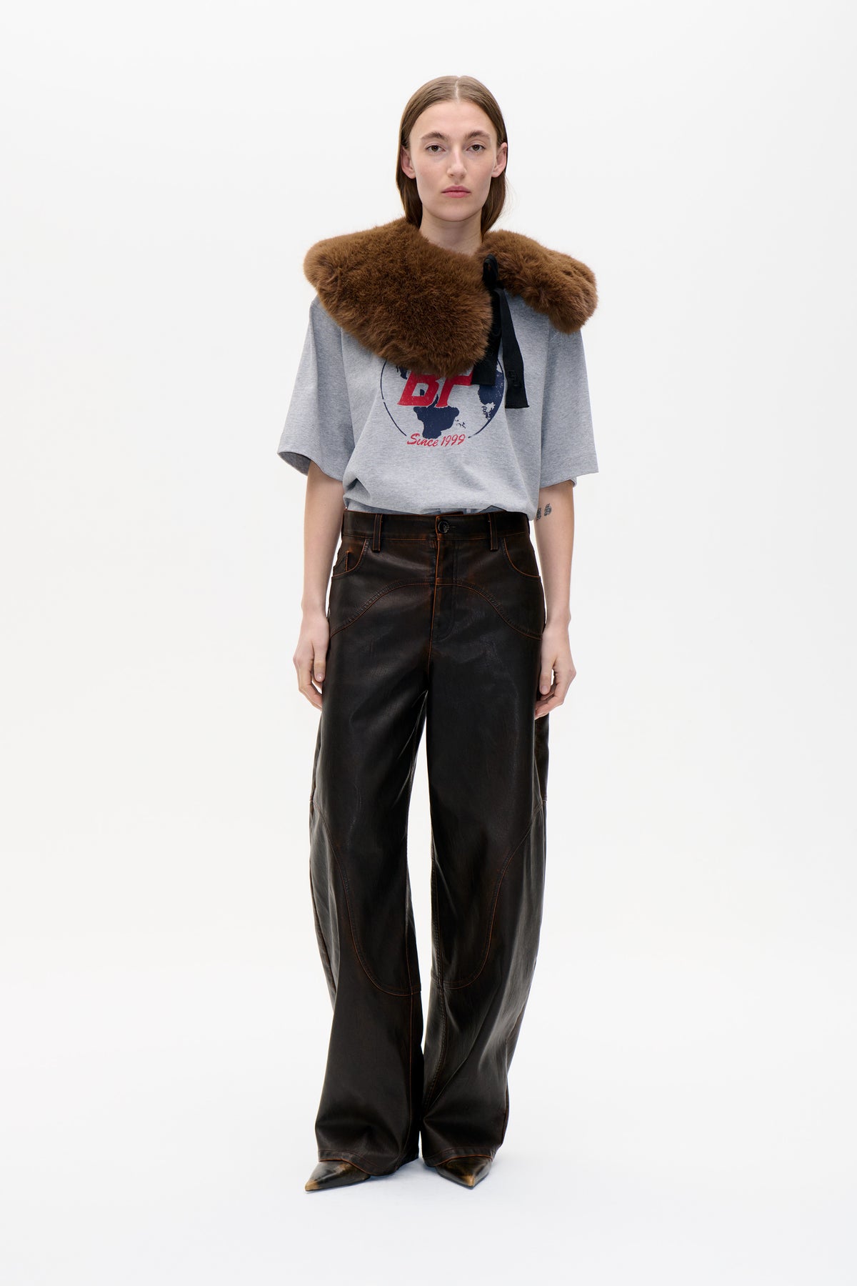 Model wearing a grey graphic T-shirt with a large brown plush shoulder wrap tied with black ribbons and dark wide-leg trousers.