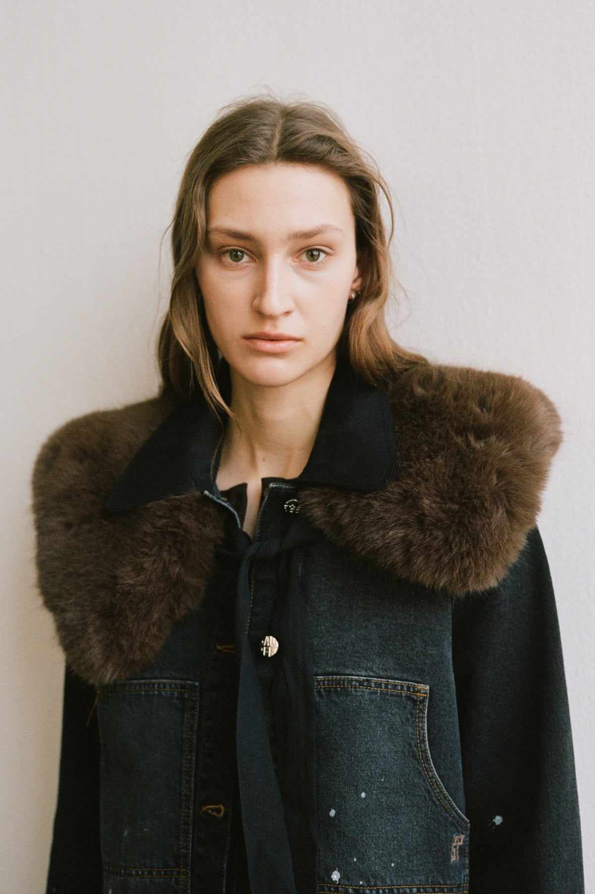 Close-up campaign portrait of a model in a dark denim jacket with contrast stitching and small white details, layered under a brown textured coat.