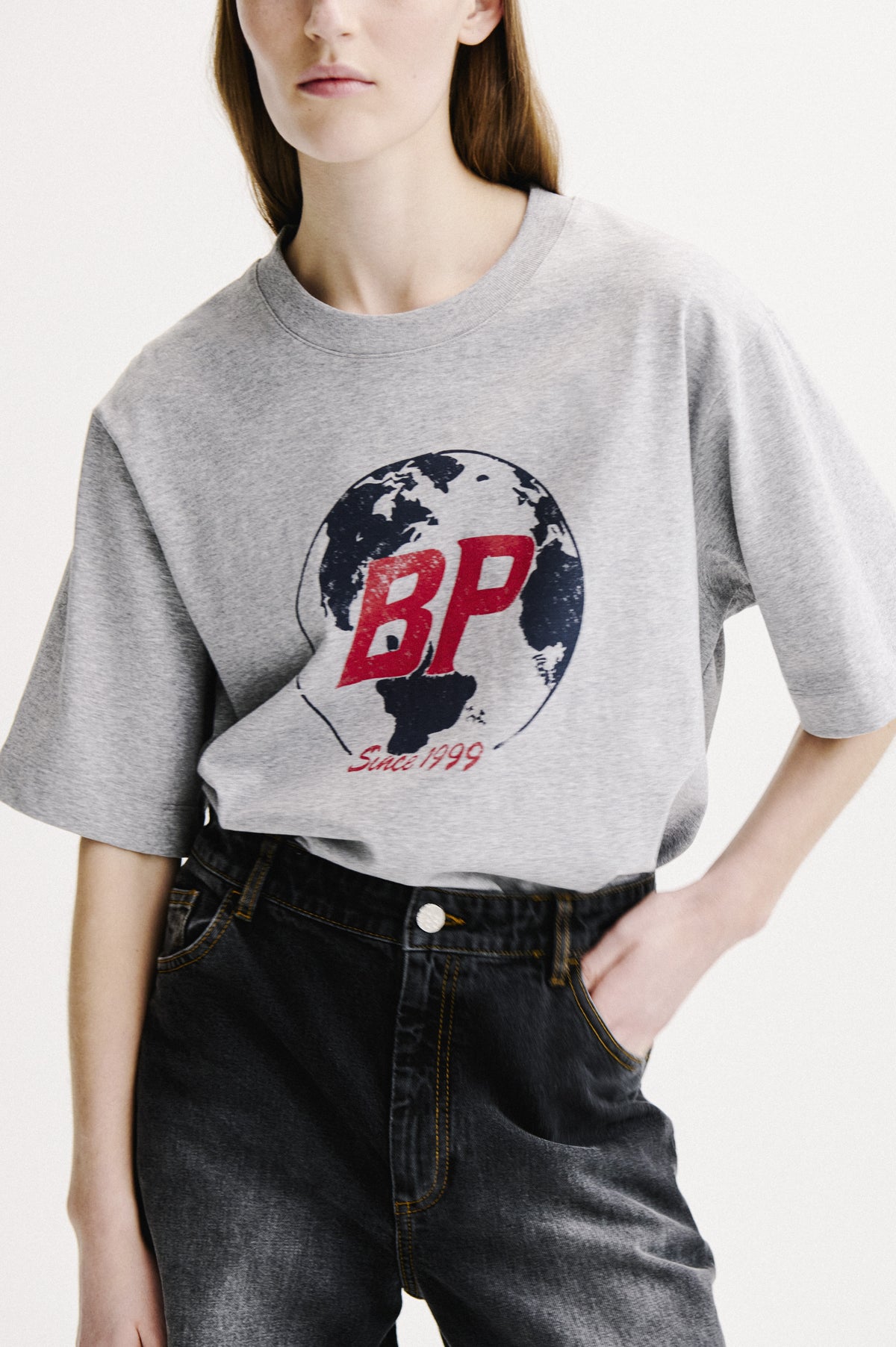 Cropped campaign portrait of a model wearing a grey marl T-shirt with a navy globe and red BP graphic, paired with dark denim jeans.
