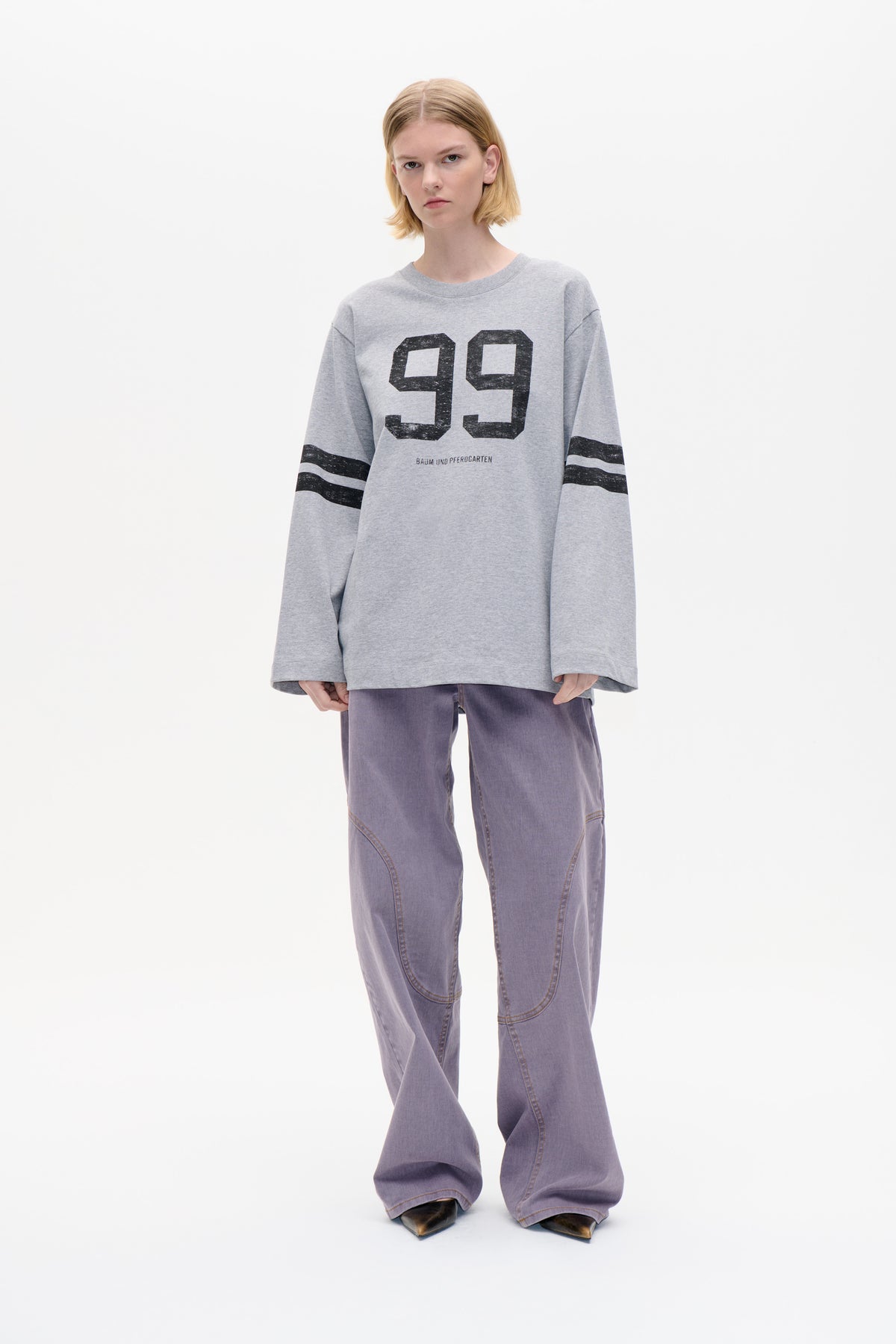 Vooraanzicht van een model in een oversized grijs sweatshirt met een grote zwarte '99'-print en gestreepte mouwen, gecombineerd met lavendelkleurige wijdvallende jeans.
