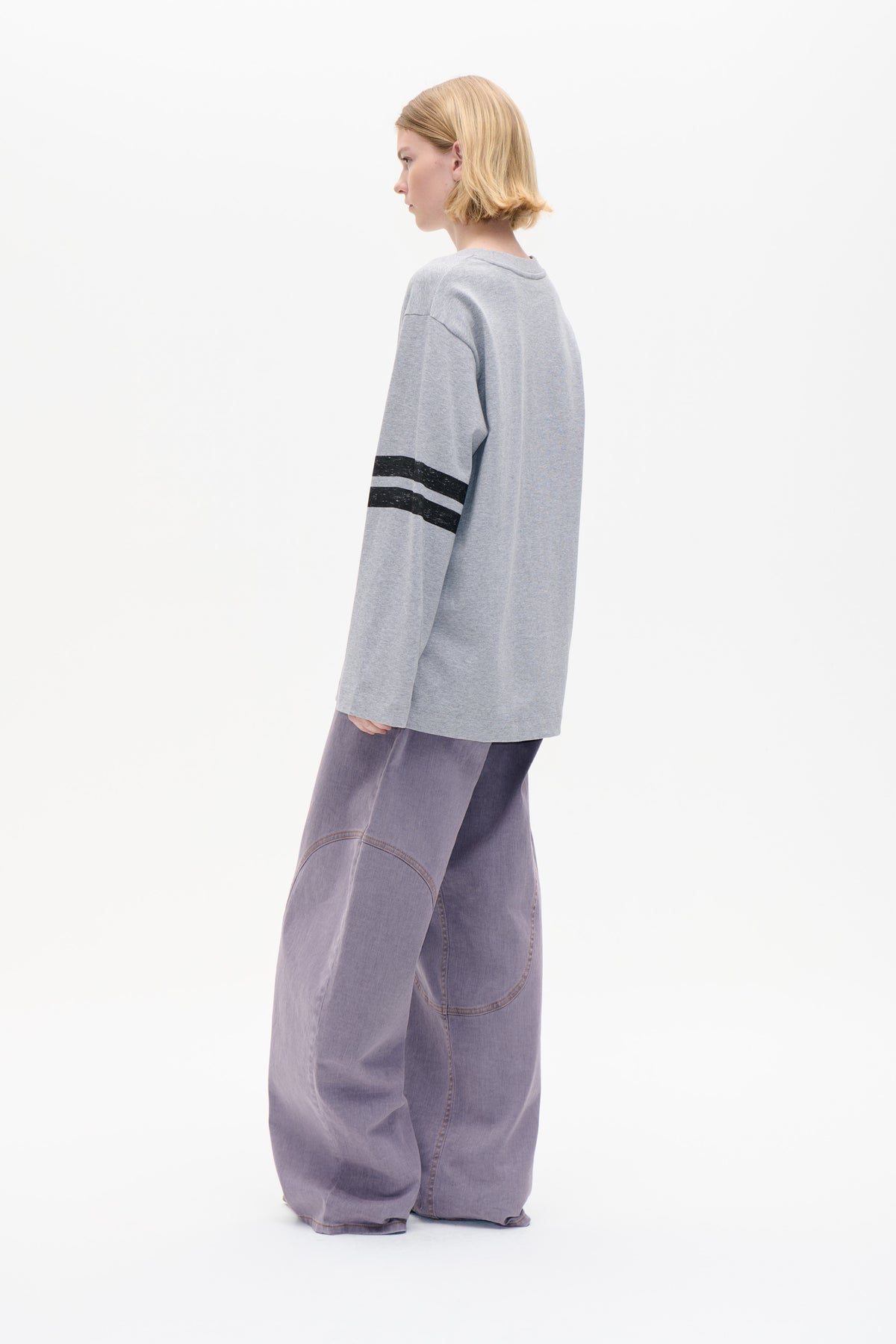 Model draagt een oversized gemêleerd grijs sweatshirt met zwarte strepen op de mouwen en een losse broek in lavendel, gefotografeerd van opzij tegen een witte achtergrond.