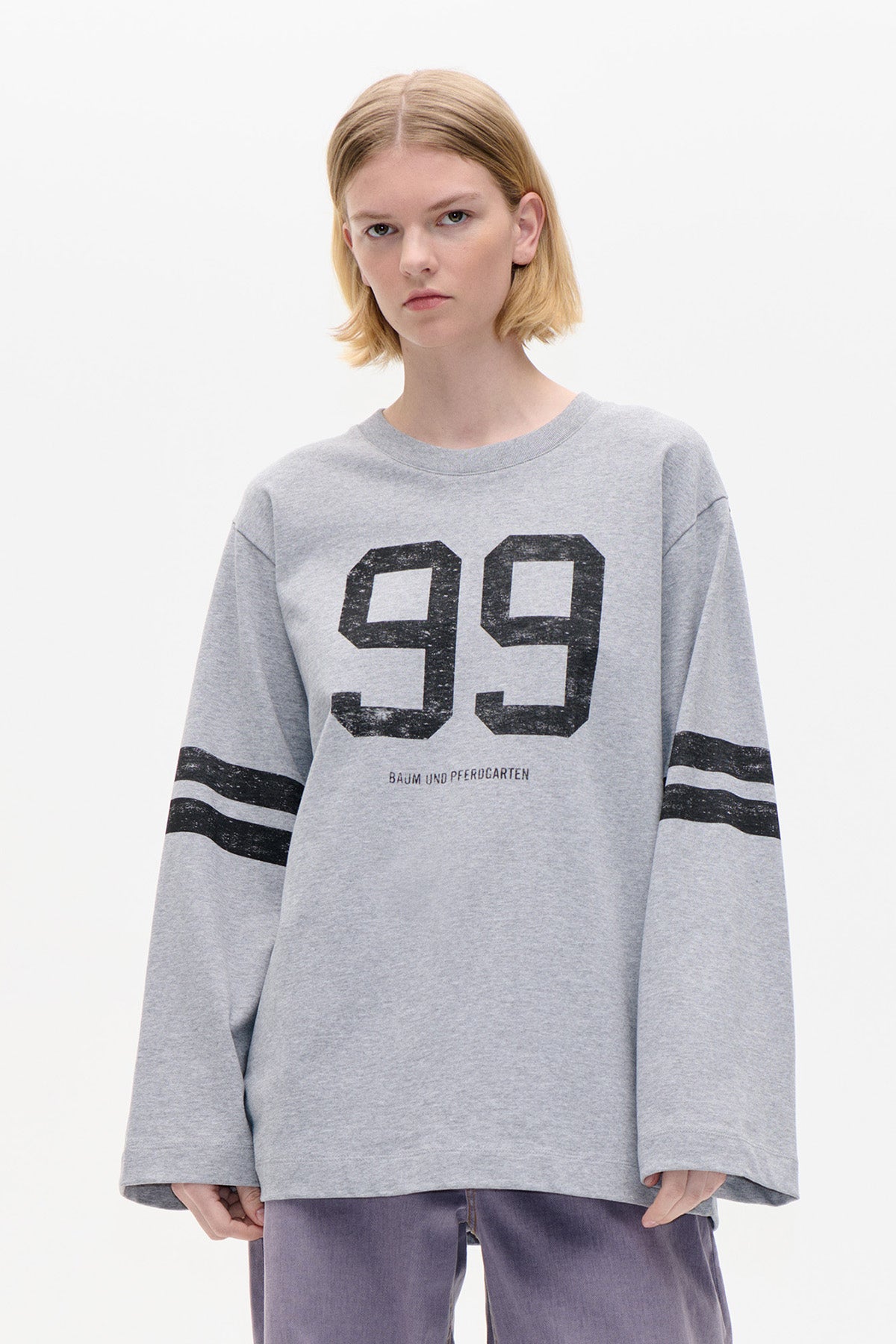 Model draagt een oversized lichtgrijze sweatshirt met zwarte '99'-print op de borst en dubbele mouwstrepen, gefotografeerd van voren.