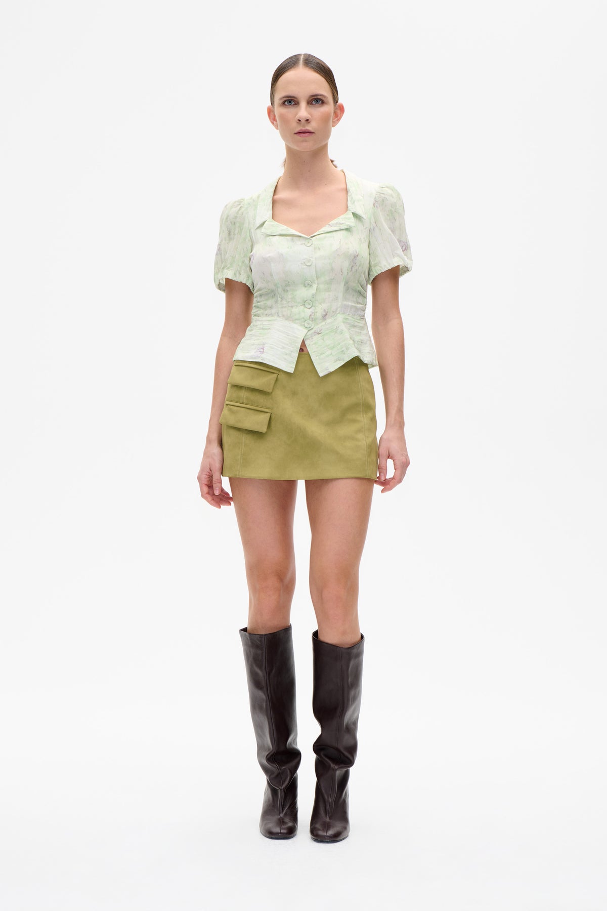 Vorderansicht eines Models, das eine blassgrün-weiß gemusterte Bluse mit abstraktem Print, Puffärmeln und Knopfleiste trägt, kombiniert mit einem olivgrünen Minirock.
