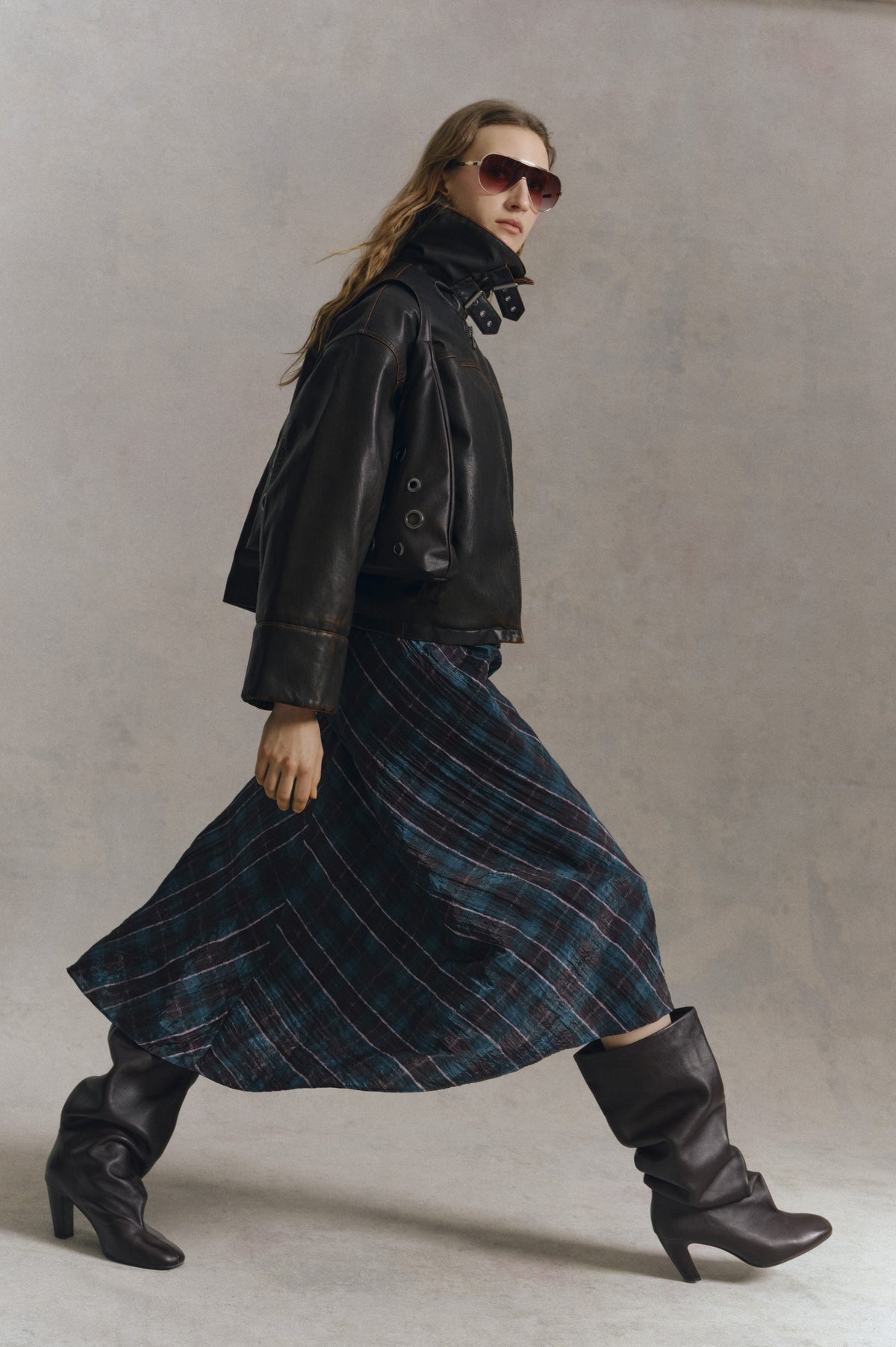 Model wearing a black leather jacket with oversized buckled collar, flowing dark teal and purple striped skirt, sunglasses and tall black boots.