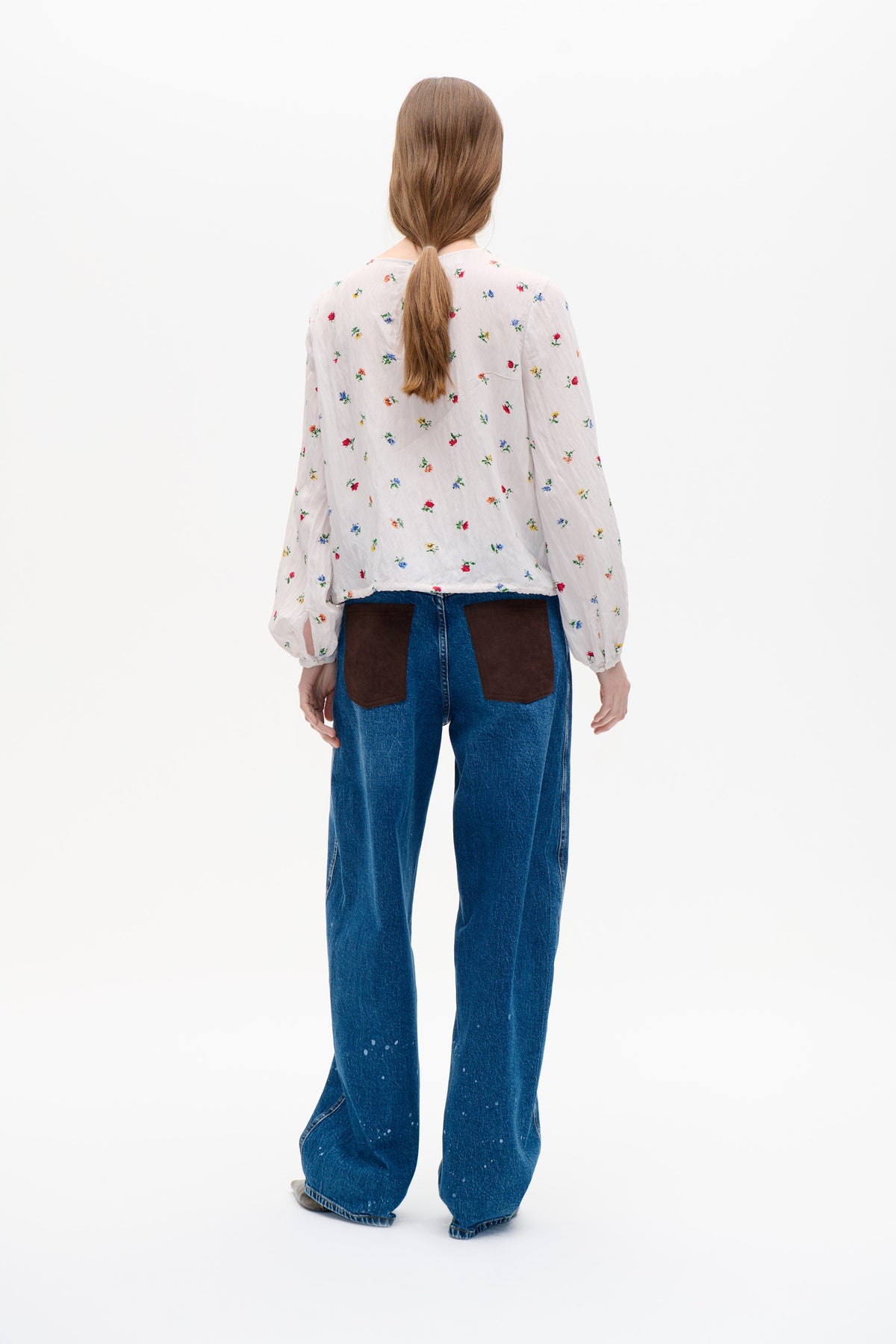 Back view of a model wearing a white blouse with scattered multicolour floral print and voluminous sleeves, paired with wide-leg blue jeans.