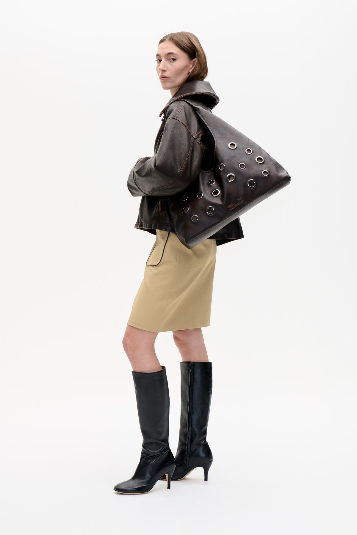 Model in a dark brown oversized jacket, carrying a matching grommeted tote, with a beige skirt and black knee-high boots.