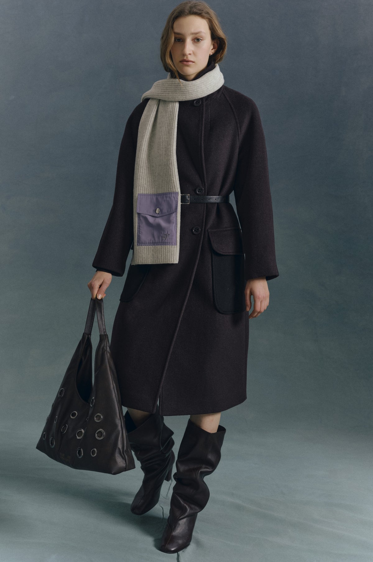 A model wears a dark button-front belted coat and cream knit scarf with a purple pocket, carrying a black eyelet tote.