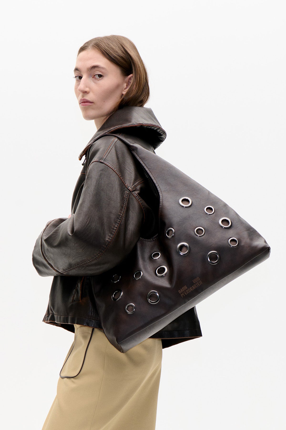 Model in a dark leather-look jacket and beige skirt carries an oversized dark brown tote with metal eyelets and Baum und Pferdgarten lettering.