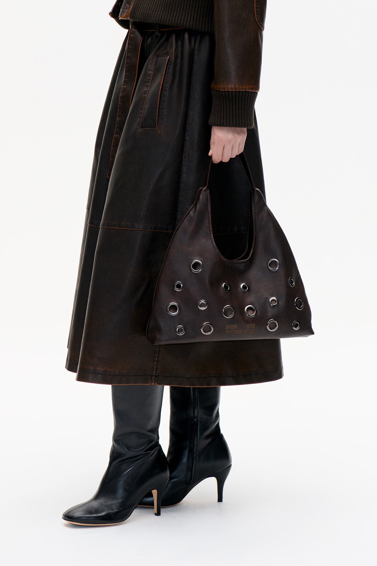Dark brown tote bag with silver-tone eyelets, carried beside a black skirt and knee-high heeled boots.