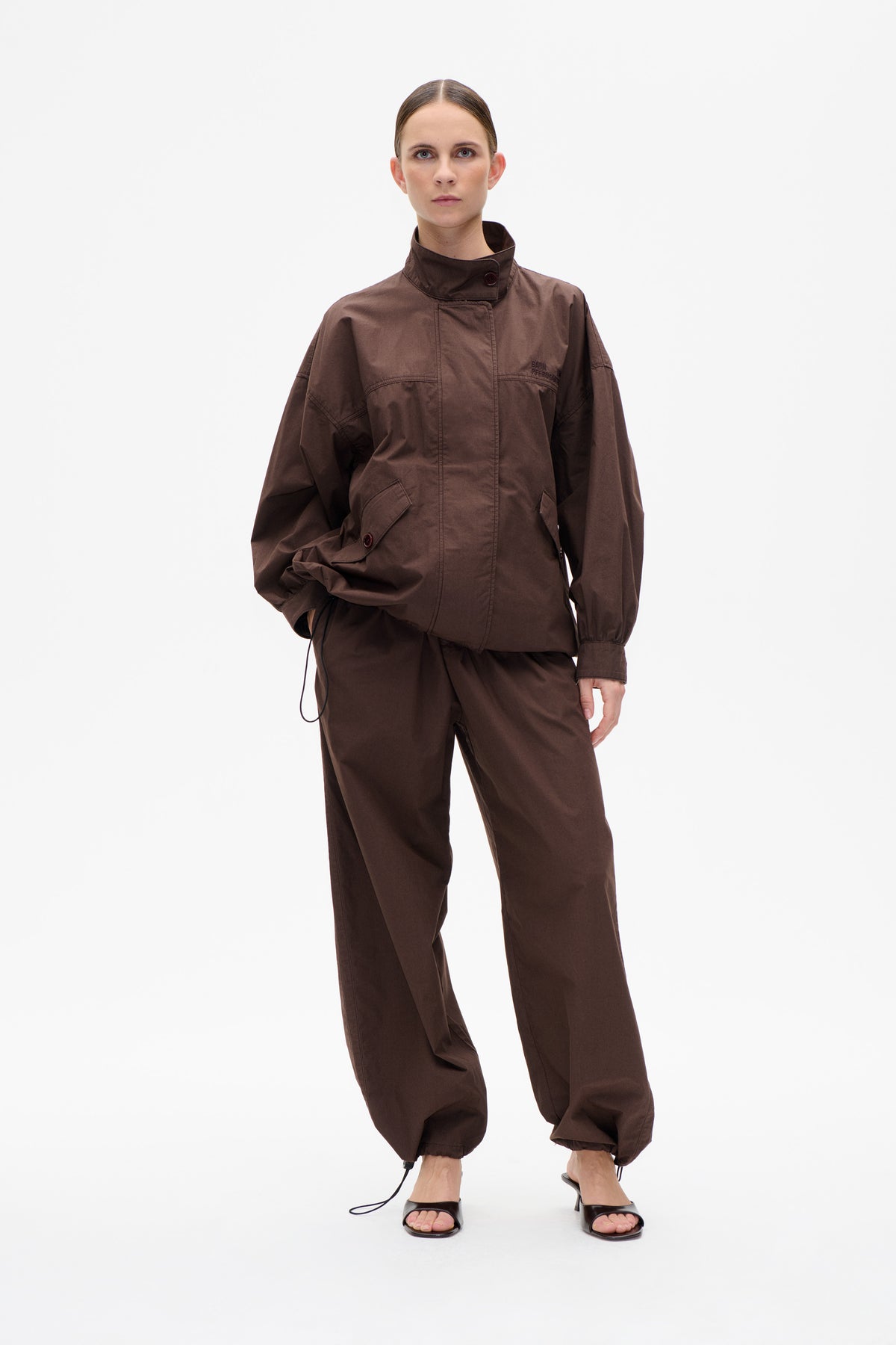 Model wearing a dark brown jacket with a high buttoned collar and large pockets, paired with matching drawstring-hem trousers.
