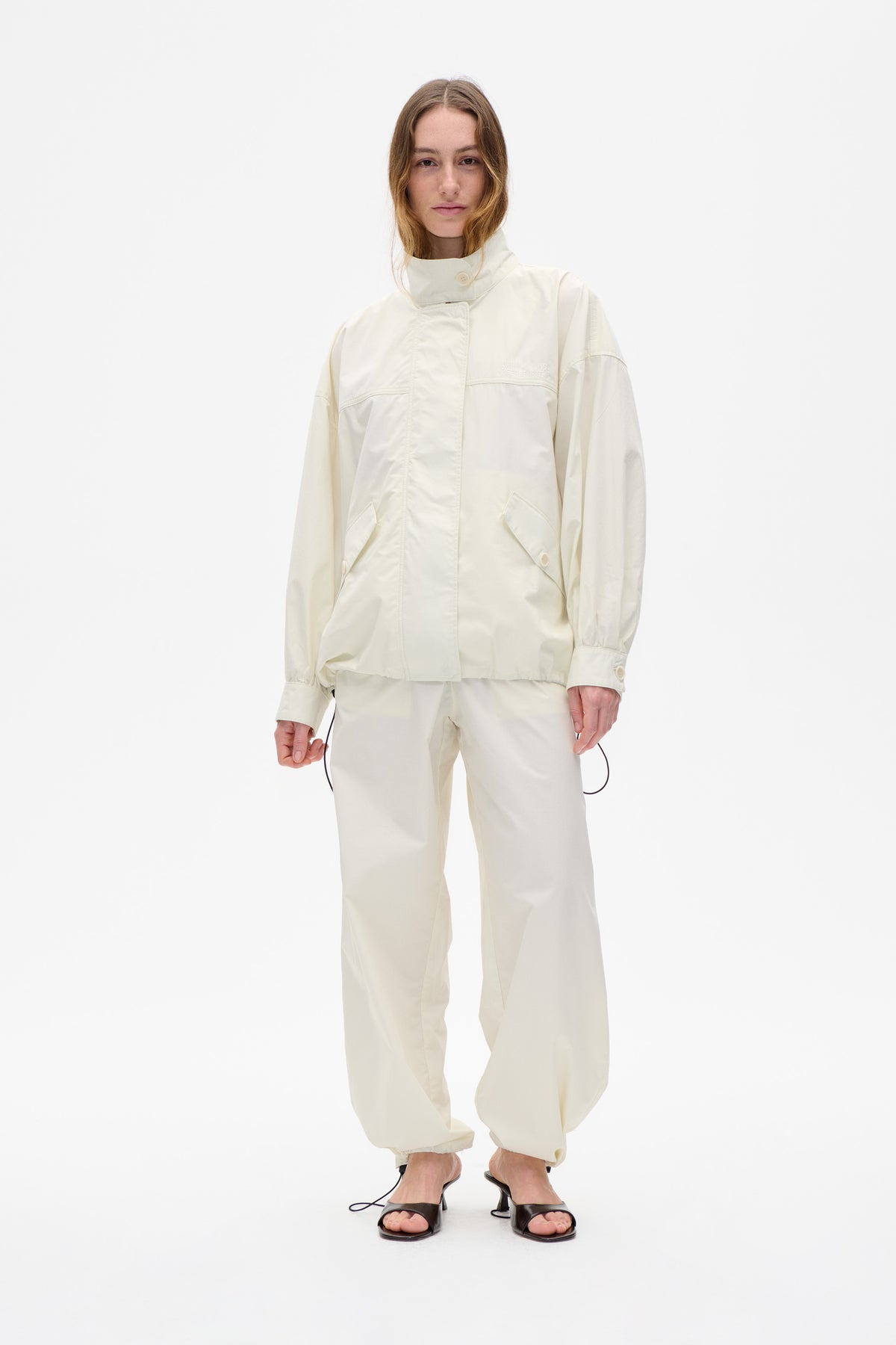 Model wearing an off-white high-collar jacket with button fastening and angled pockets, paired with matching trousers against a white backdrop.