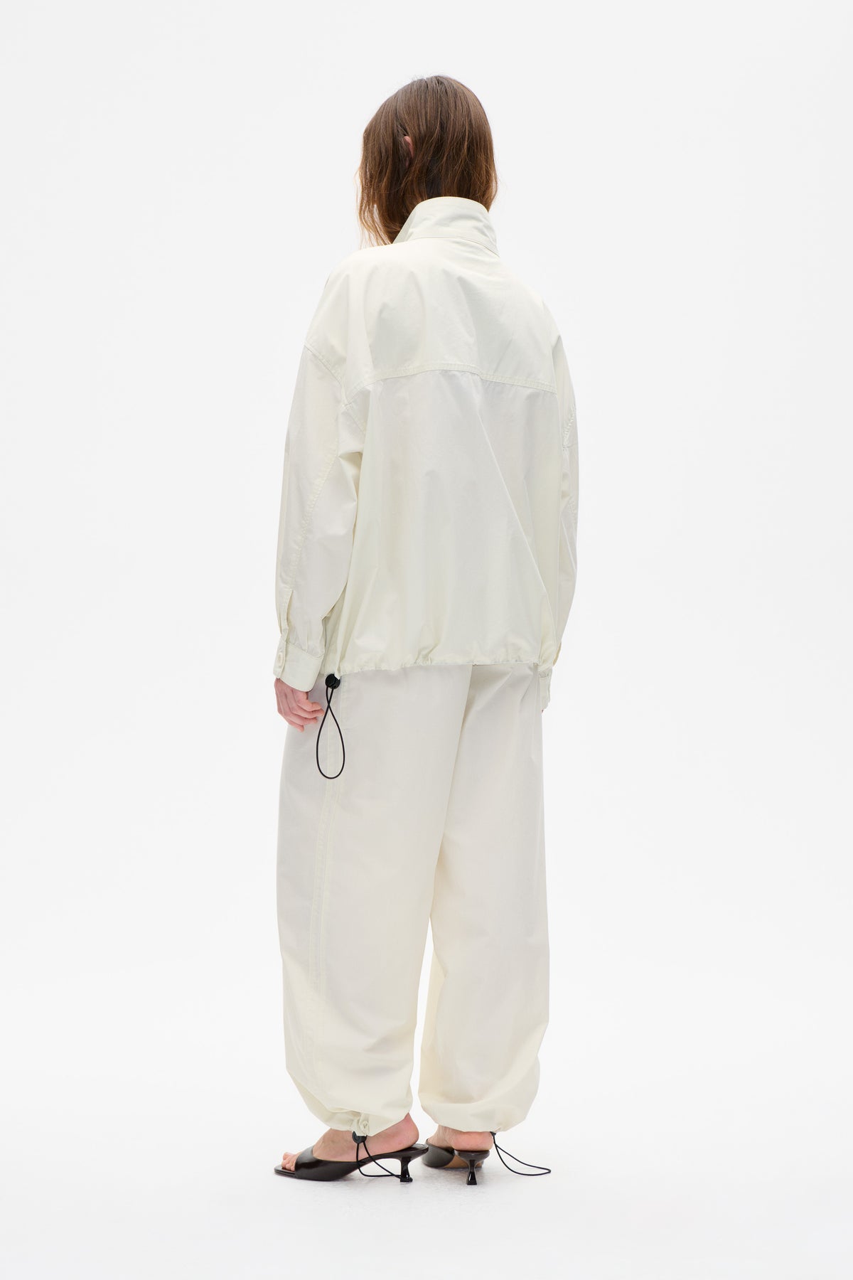 Back view of a model wearing an off-white jacket with a high collar and curved yoke seam, paired with matching trousers.