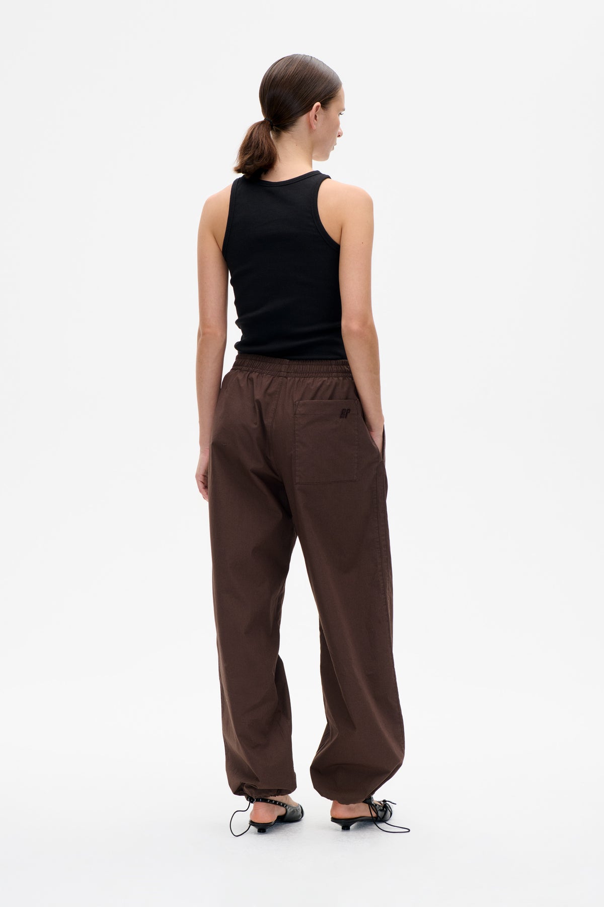 Back view of a model wearing a black sleeveless top and loose brown trousers with an elastic waist and back pocket.