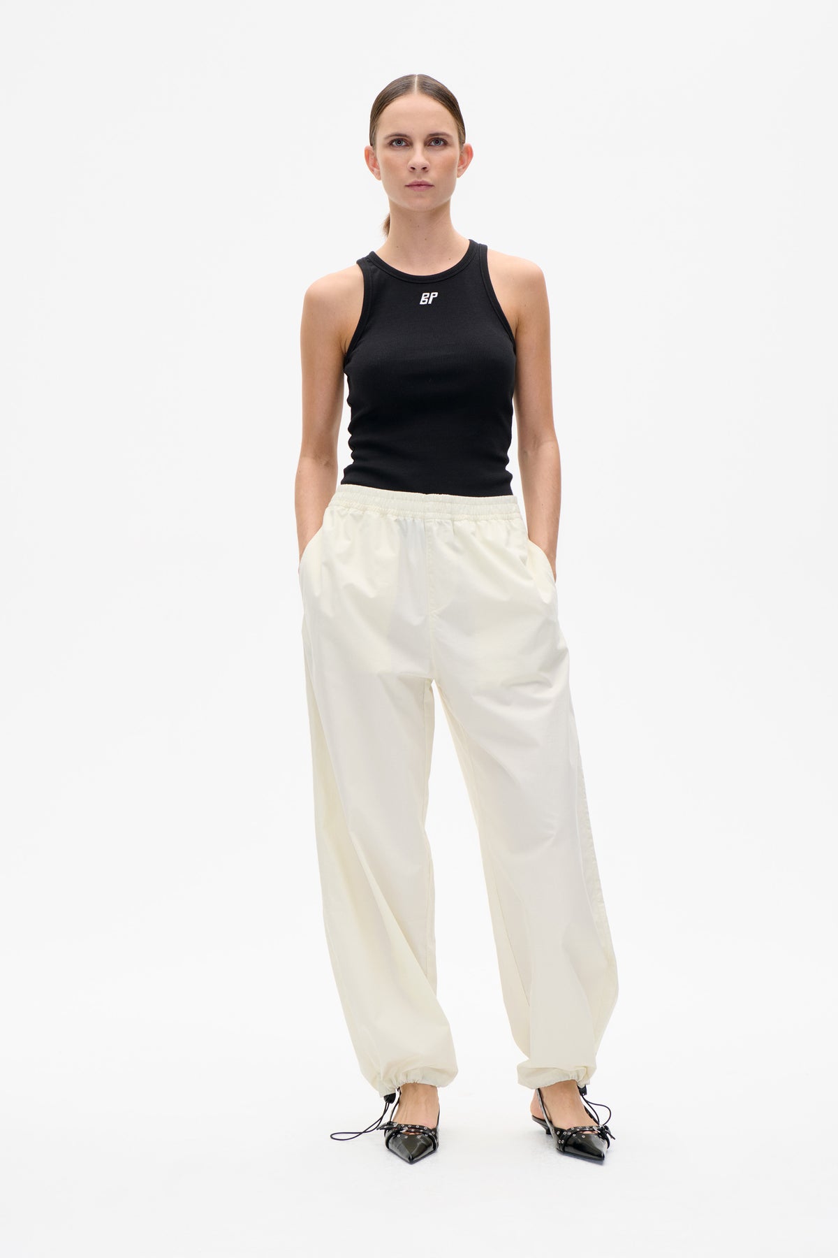 Model, von vorn gezeigt, trägt ein schwarzes, ärmelloses Ripp-Top mit kleinem weißen Monogramm und eine weite elfenbeinfarbene Hose.