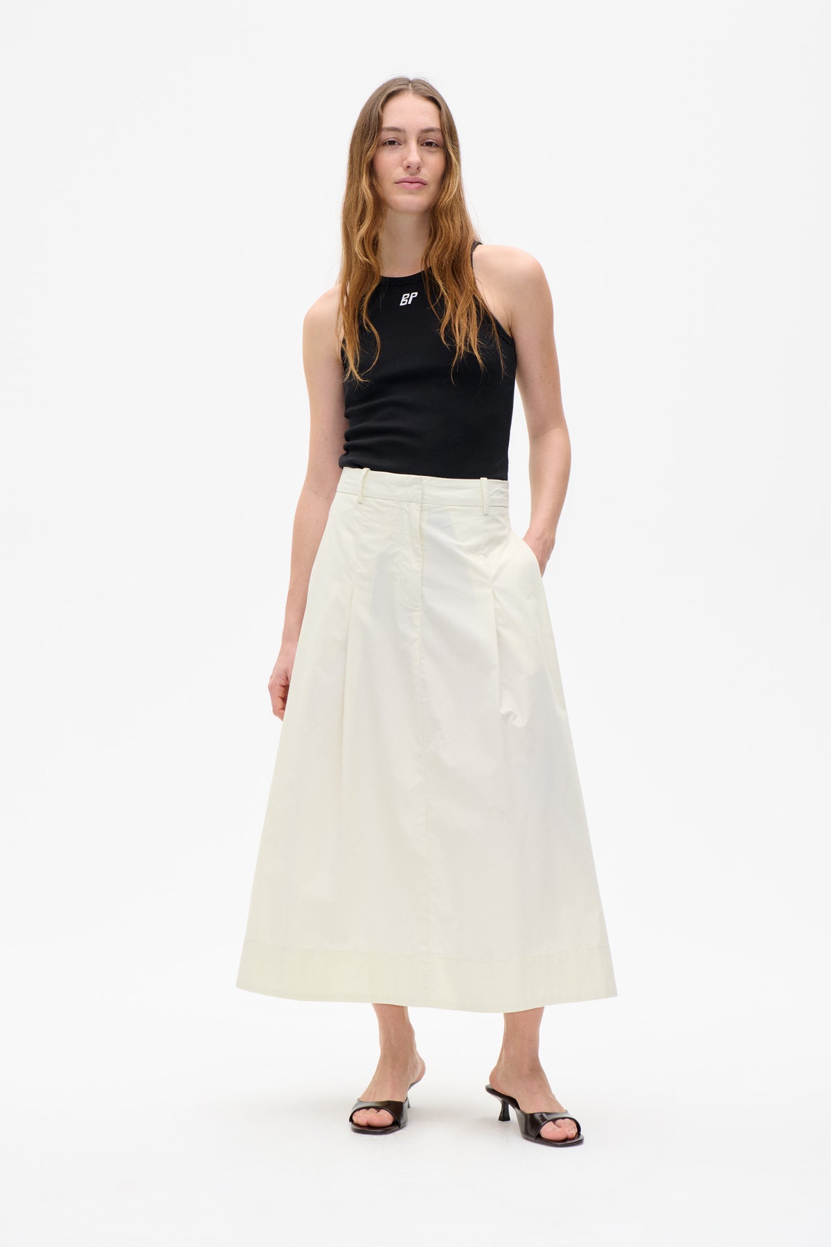 Vorderansicht eines Models, das ein schwarzes, ärmelloses, geripptes Top mit einem kleinen weißen Brustlogo und einen elfenbeinfarbenen, hochgeschnittenen Midirock trägt.