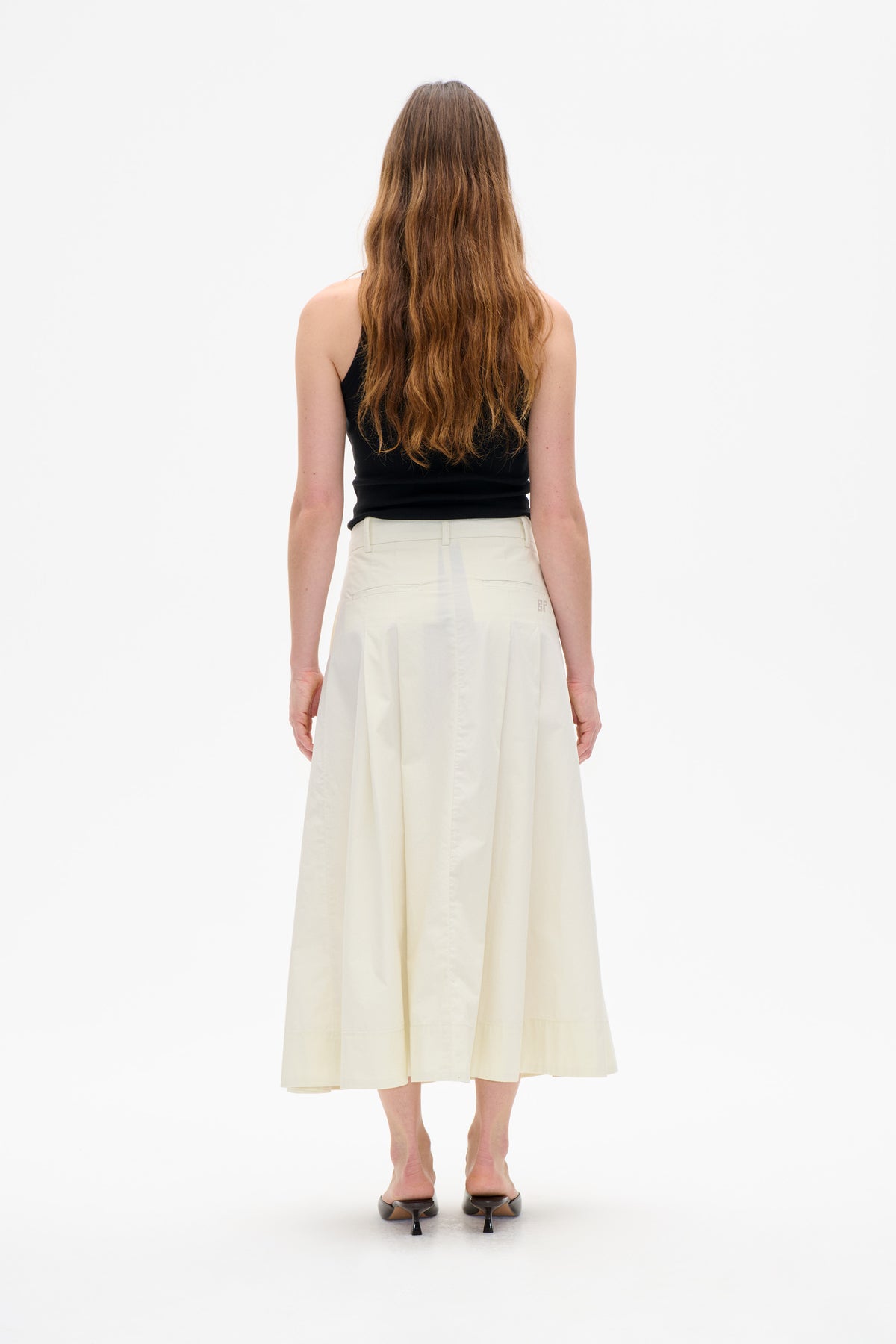 Rear view of a model wearing an ivory panelled midi skirt with belt loops and back welt pockets, paired with a black sleeveless top.