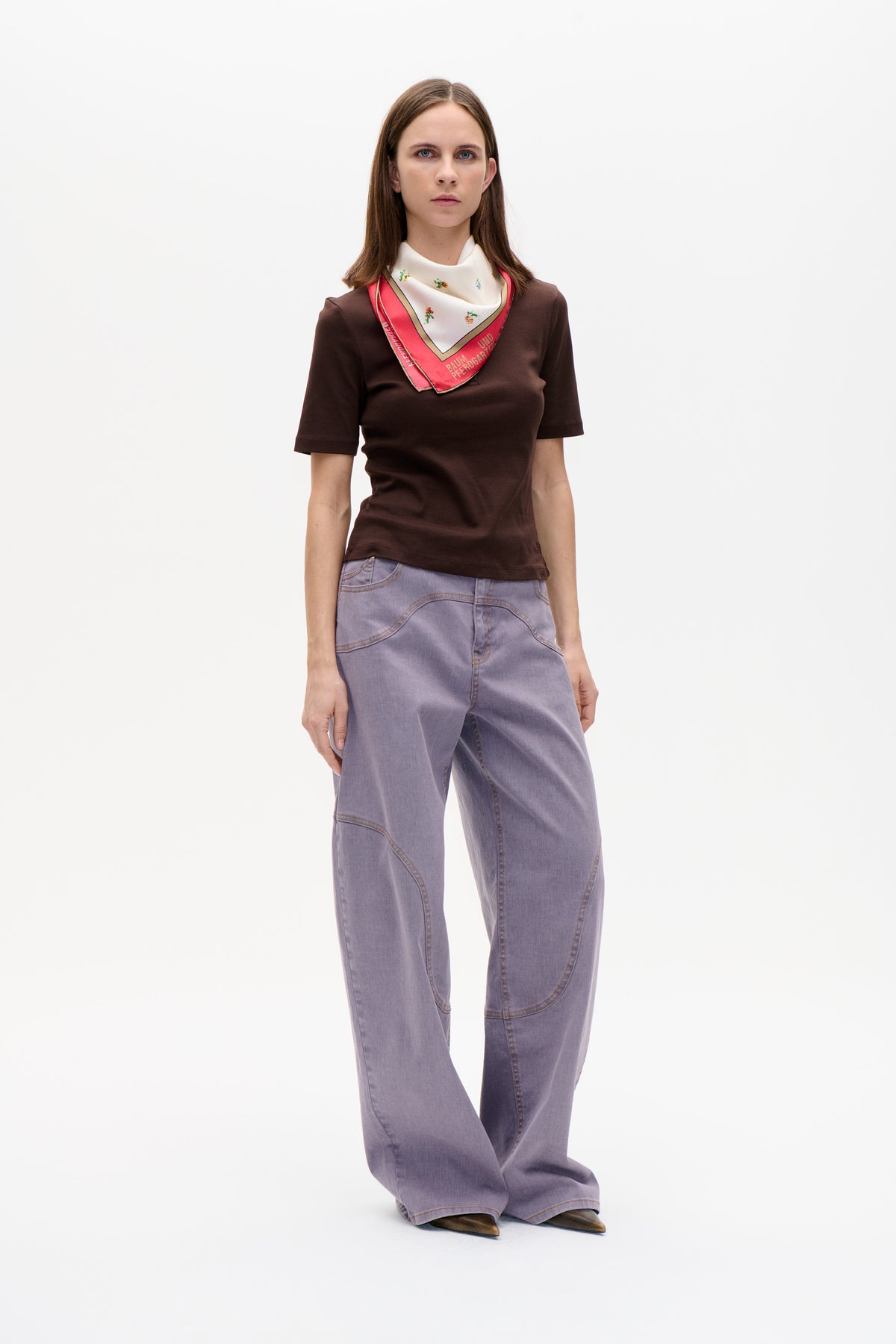 Model trägt ein dunkelbraunes Top mit kurzen Ärmeln, fliederfarbene Jeans mit weitem Bein und einen cremefarbenen, geblümten Schal mit korallenfarbenem Rand, auf dem Baum und Pferdgarten steht.