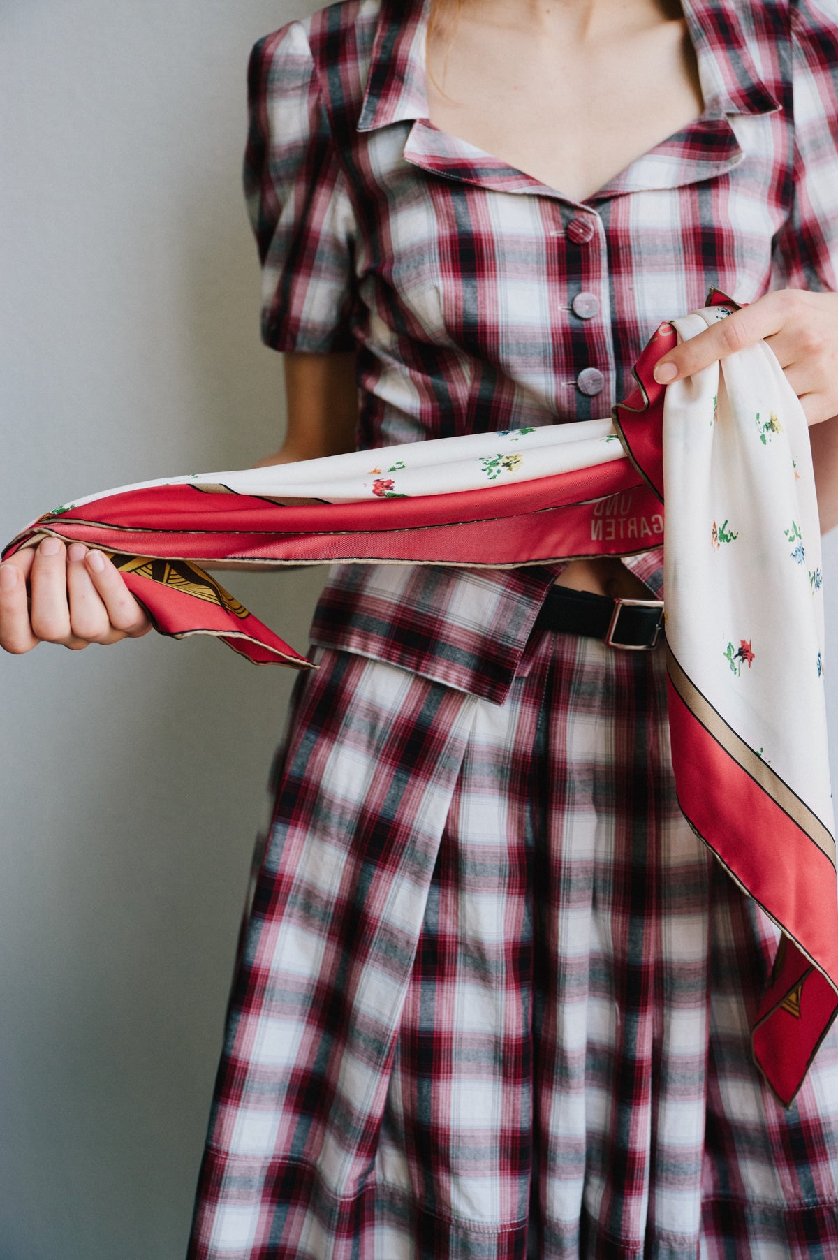 Kampagnen-Nahaufnahme eines Models, das ein kariertes Kleid in Rot, Weiß und Anthrazit mit durchgehender Knopfleiste und dunklem Gürtel trägt und bedruckte Schals hält.