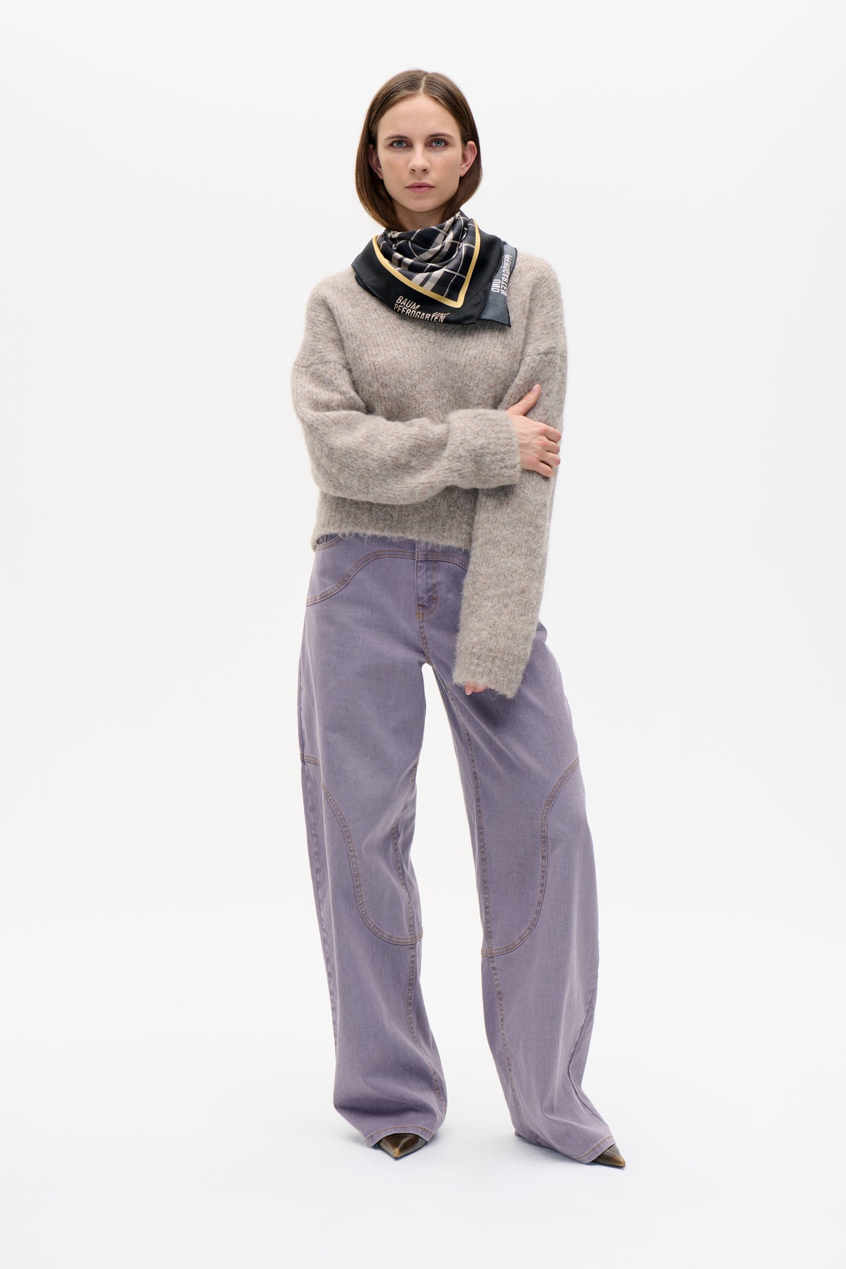 Model draagt een pluizige beige trui, lila wijde jeans en een zwarte geruite sjaal met gele bies en merkletters.
