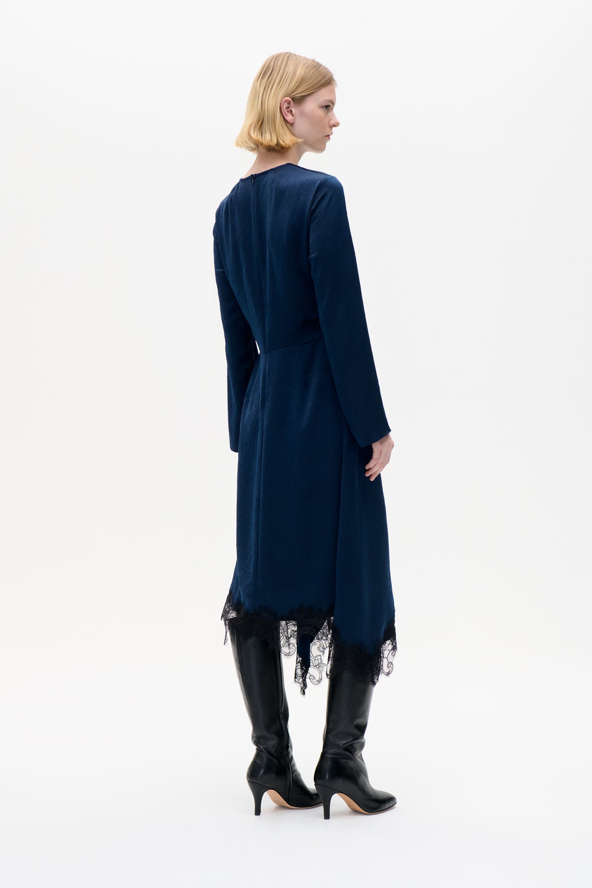 Model trägt ein marineblaues, langärmliges Kleid mit strukturierter Oberfläche und schwarzem Spitzenbesatz am Saum, von hinten vor weißem Hintergrund gezeigt.