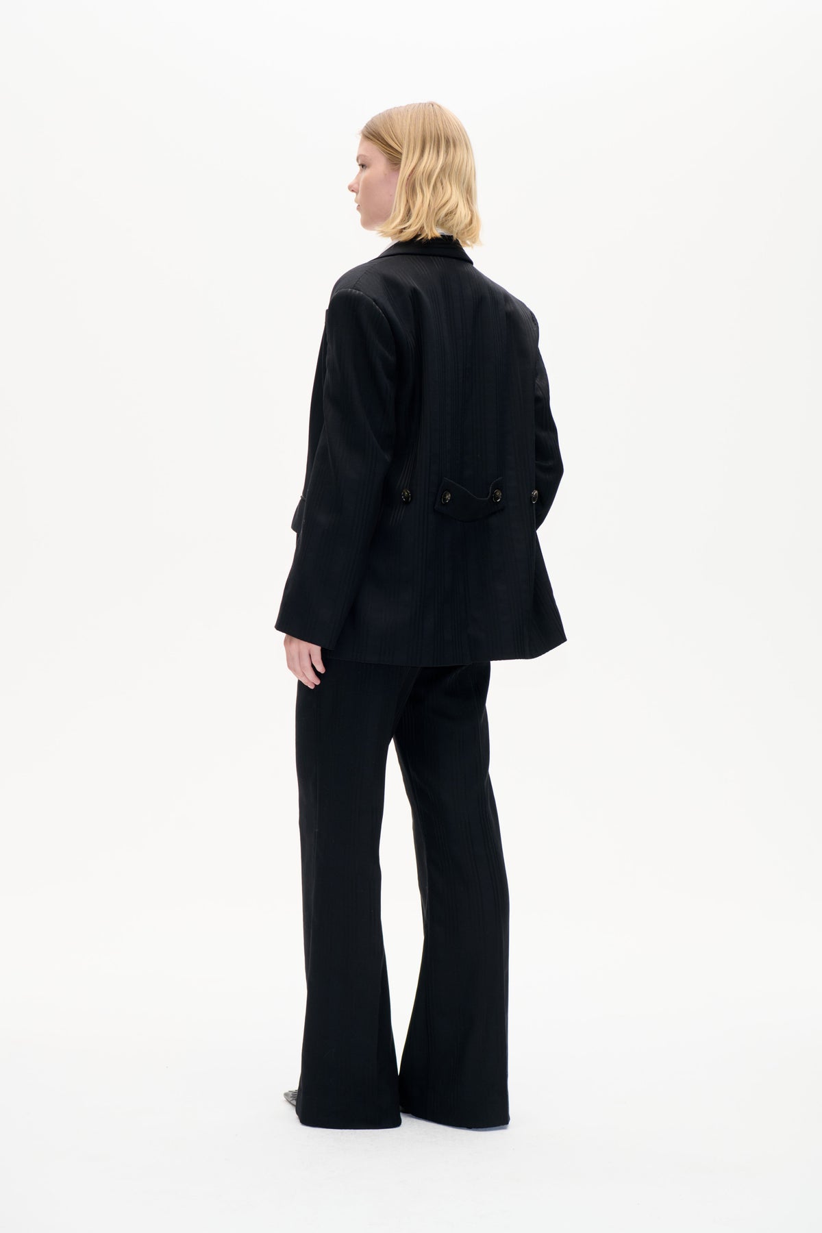 Mannequin portant un blazer noir à fines rayures, avec une patte arrière boutonnée à la taille et un pantalon à jambes larges, vu de profil et de dos.