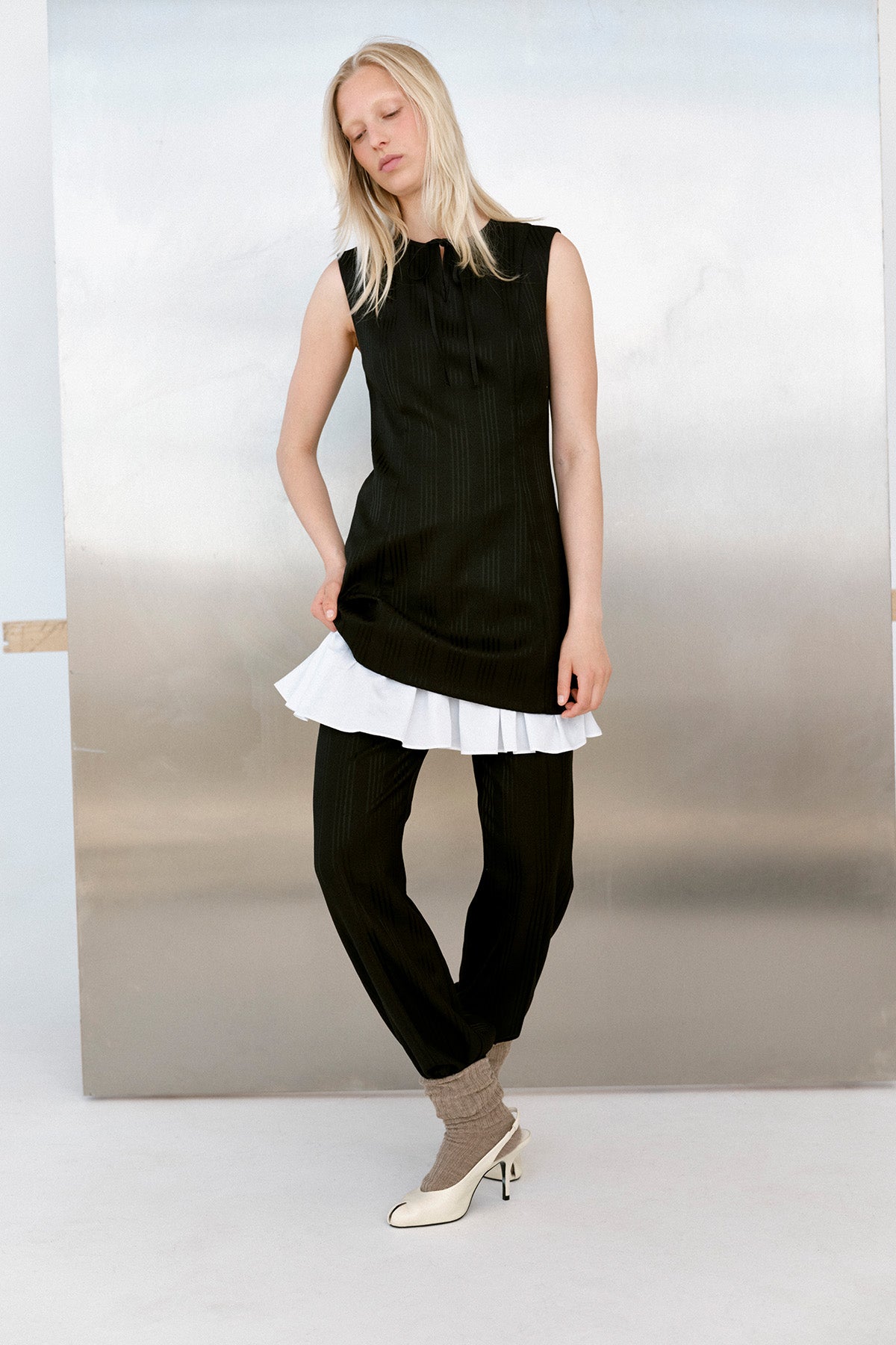 Mannequin portant une tunique noire sans manches à fines rayures verticales et encolure à nouer, superposée à un ourlet volanté blanc et portée avec un pantalon assorti.