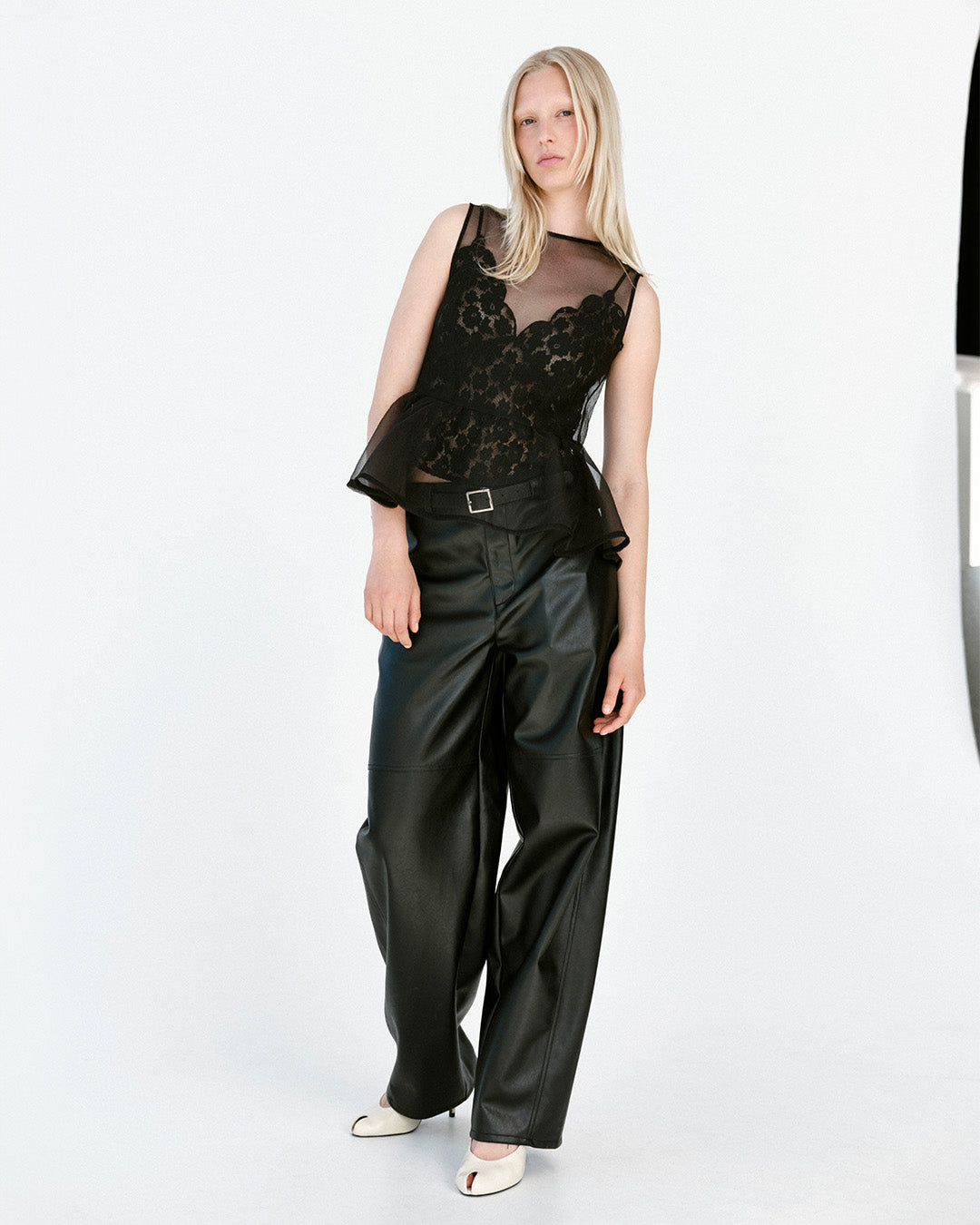 Model wearing a sheer black lace peplum top, wide-leg black leather-look trousers and white shoes against a white backdrop.