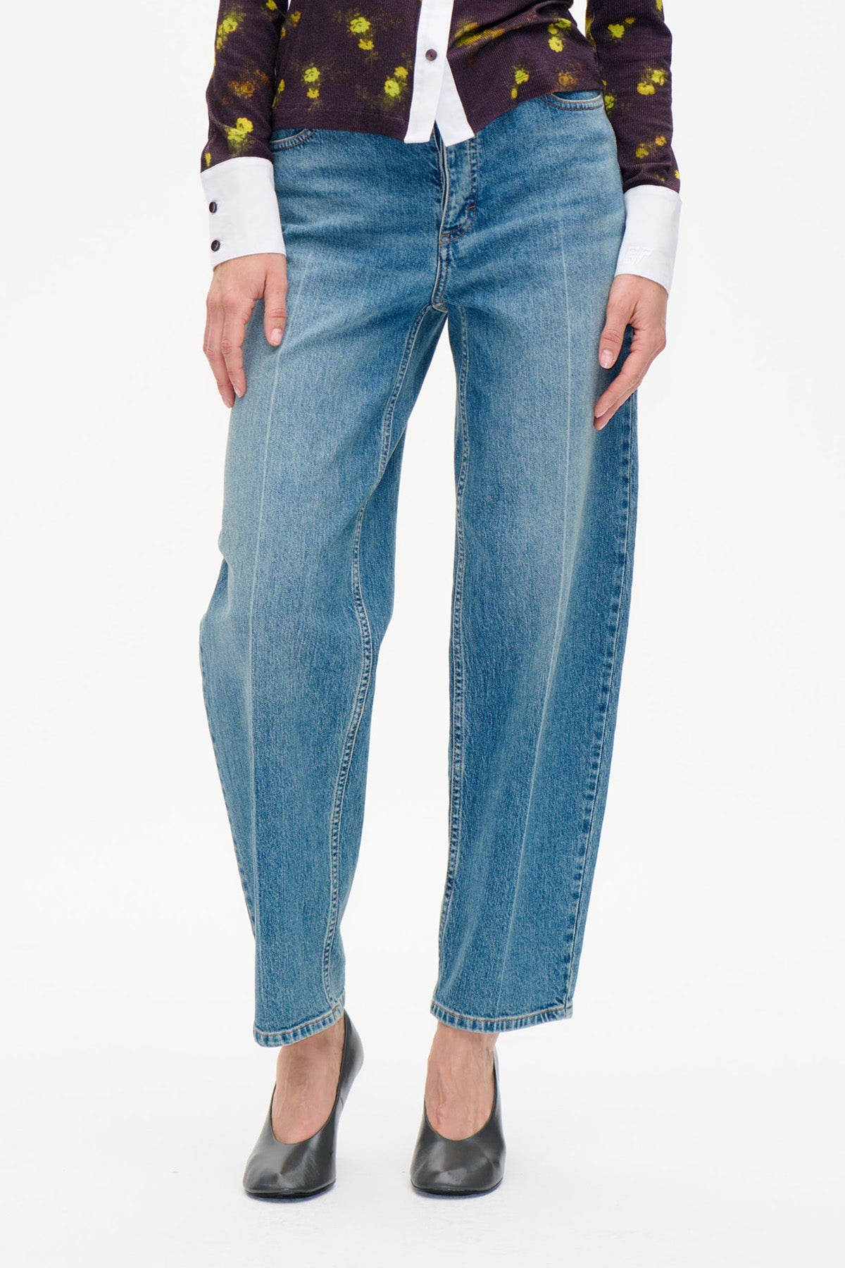 Detailaufnahme der Vorderseite einer blauen, gerade geschnittenen Jeans mit verwaschenem Finish, kombiniert mit einem dunklen, floral bedruckten Top und schwarzen Pumps.