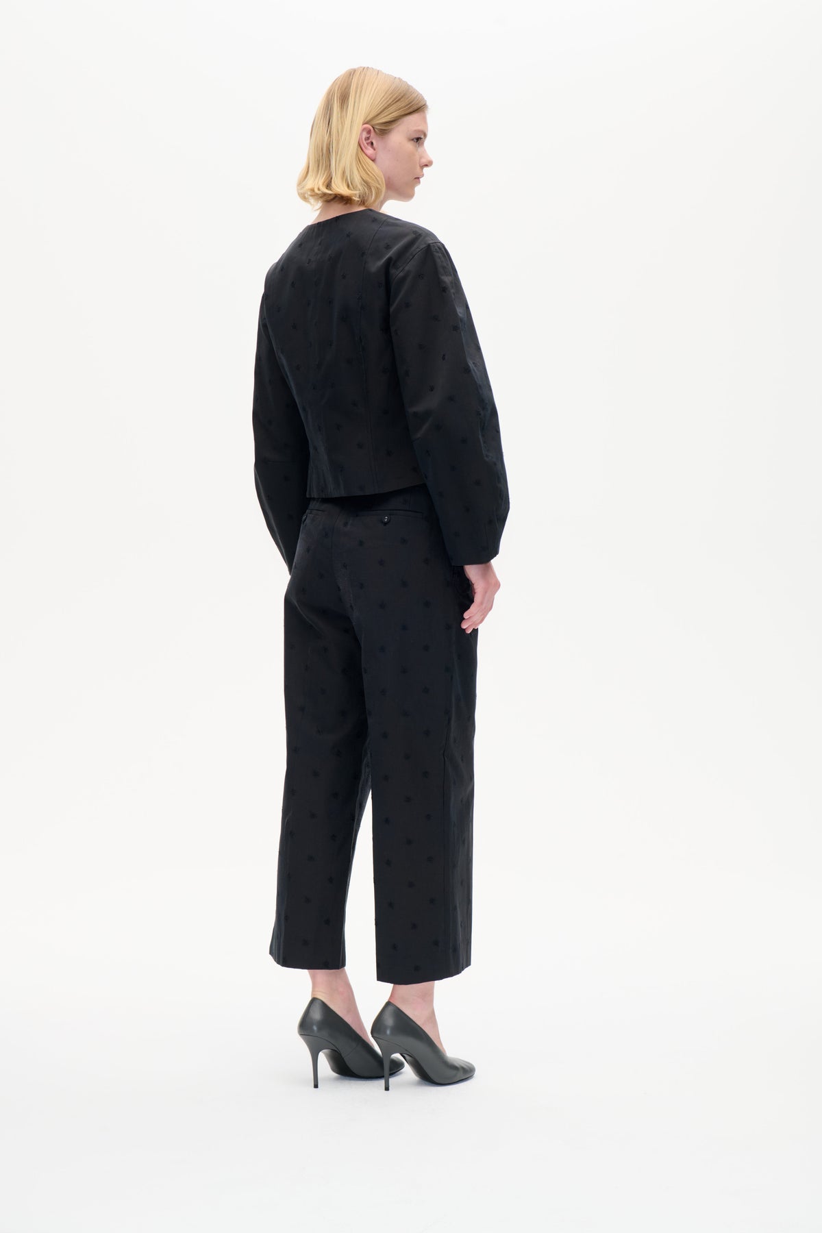 Ein Model in einer dreiviertel-Rückenansicht trägt ein schwarzes Langarmtop und gekürzte, weit geschnittene Hosen mit dezenten Ton-in-Ton-Motiven vor weißem Hintergrund.