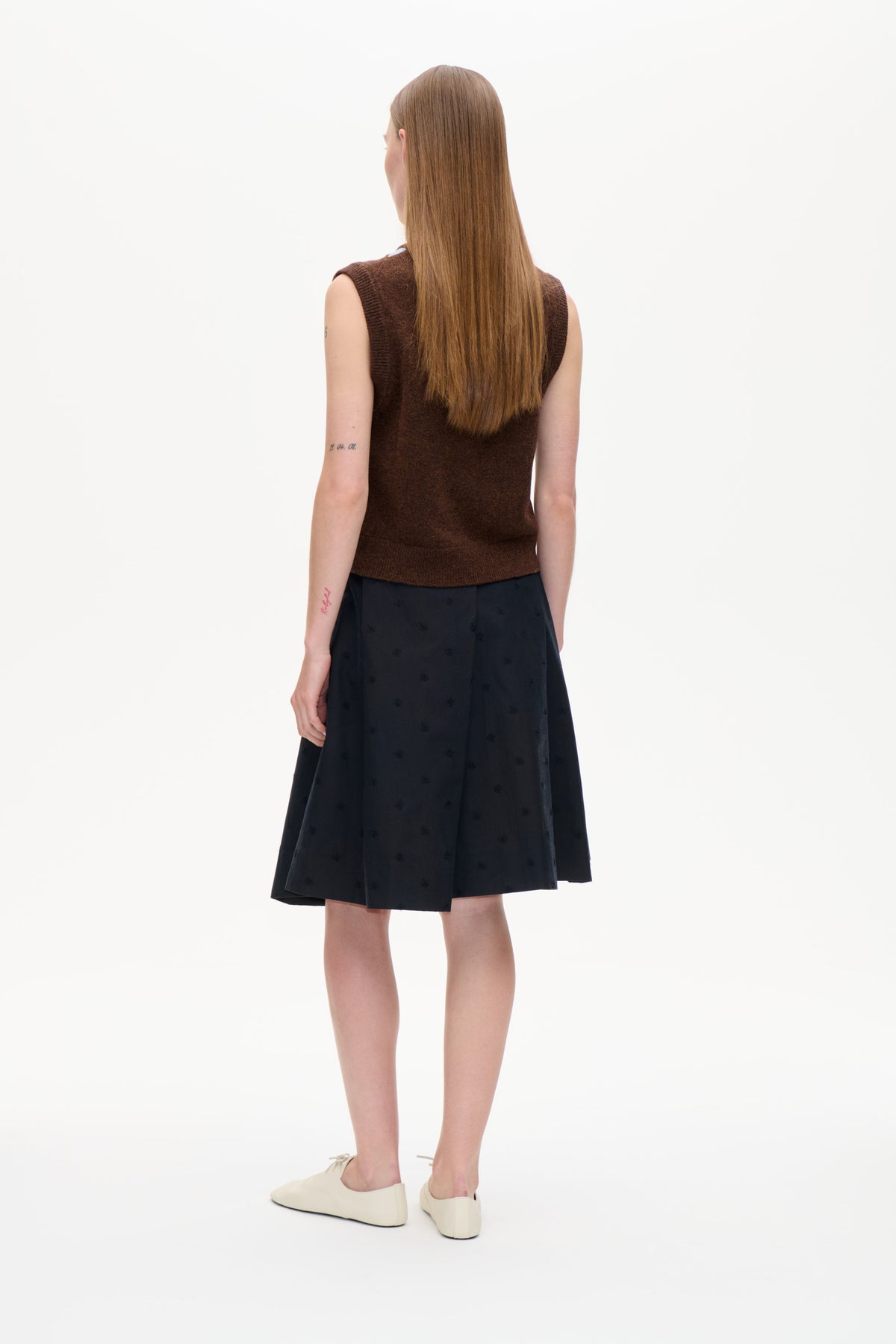 Back view of a model wearing a dark brown sleeveless knit top, a black skirt with subtle motifs, and cream flats.