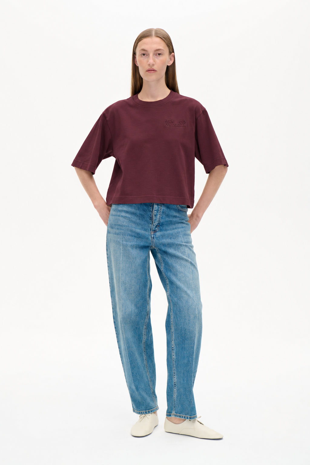 Vooraanzicht van een model dat een bordeauxkleurig T-shirt met korte mouwen en een tonaal logodetail op de borst draagt, gecombineerd met lichtblauwe jeans met patroon en wijde pijpen.