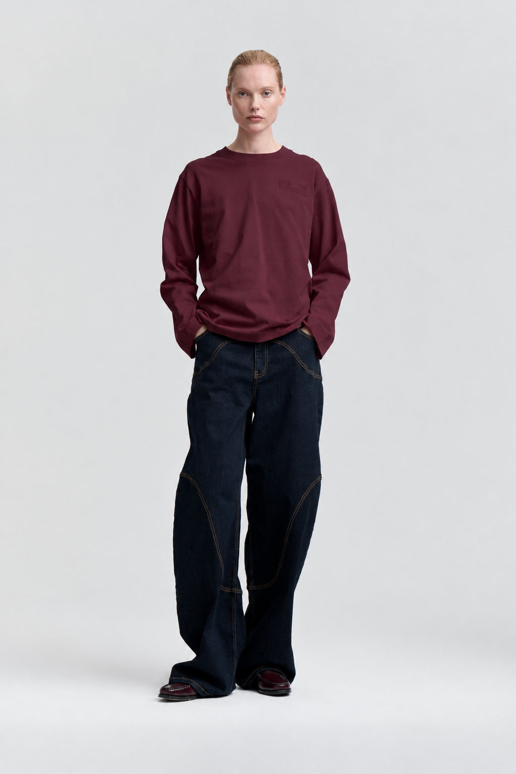 Vooraanzicht van een model dat een bordeauxrood sweatshirt met lange mouwen draagt, gecombineerd met wijde, donkerindigo jeans met contrasterende stiksels.