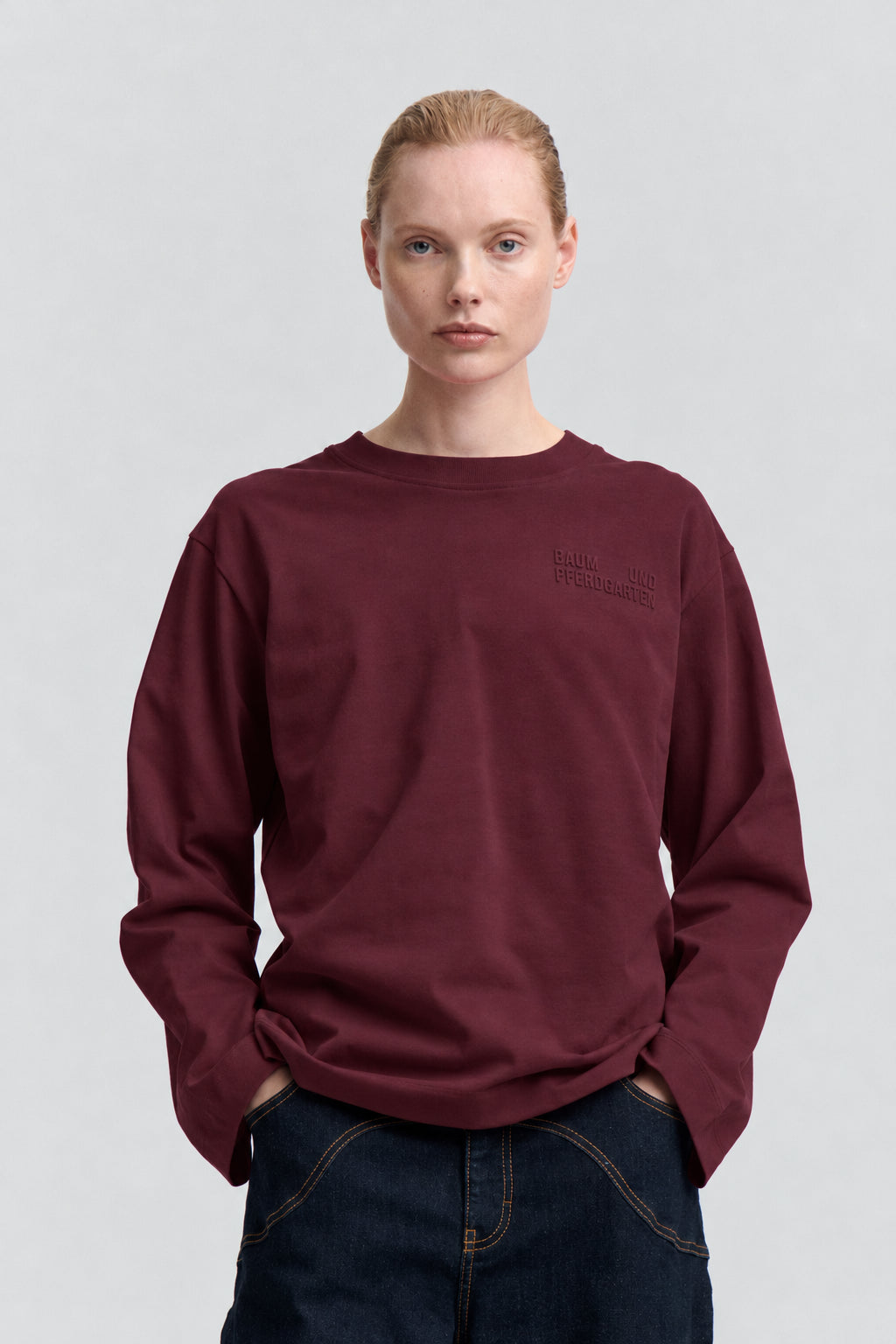 Vooraanzicht van een sweatshirt in bordeauxrood met ronde hals en lange mouwen, met subtiele tonale tekst op de borst, gestyled met donkere jeans.