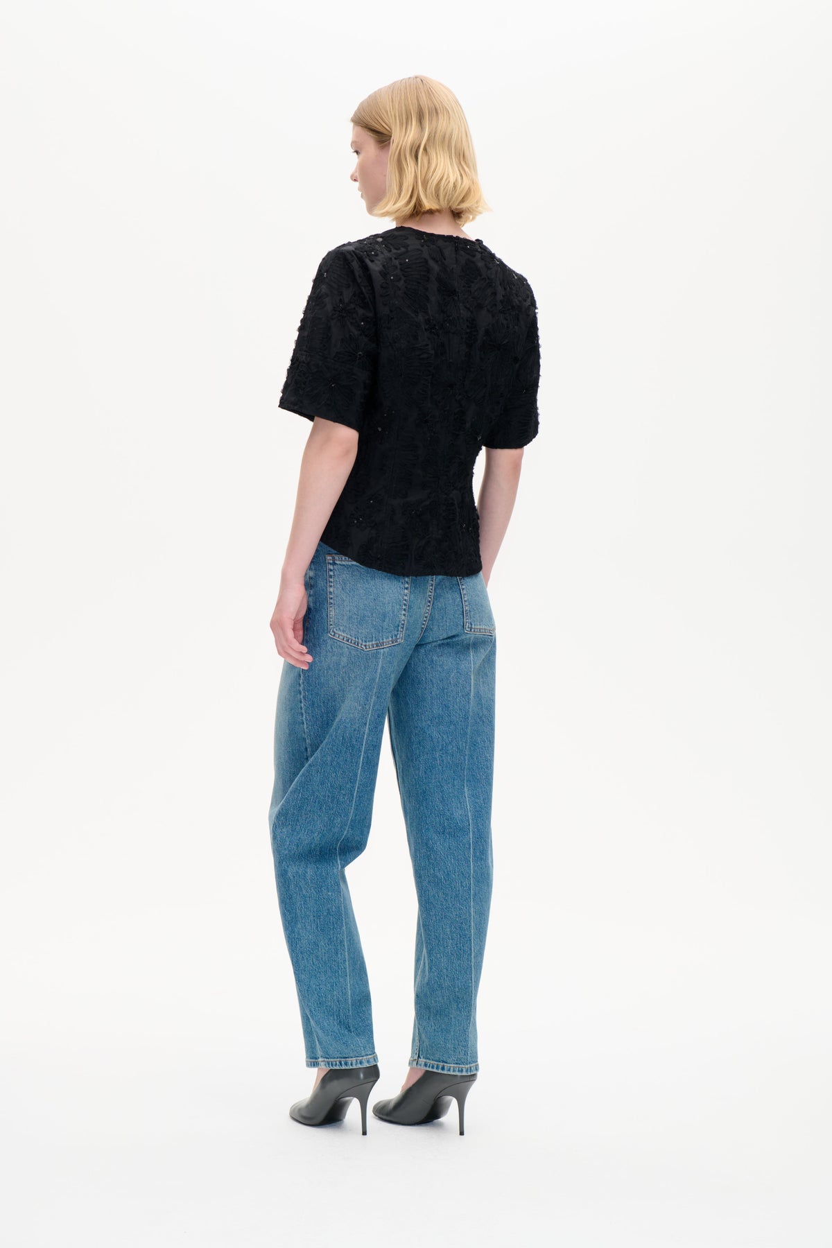 Model viewed from behind wearing a black short-sleeved top with textured floral embellishment and blue jeans.