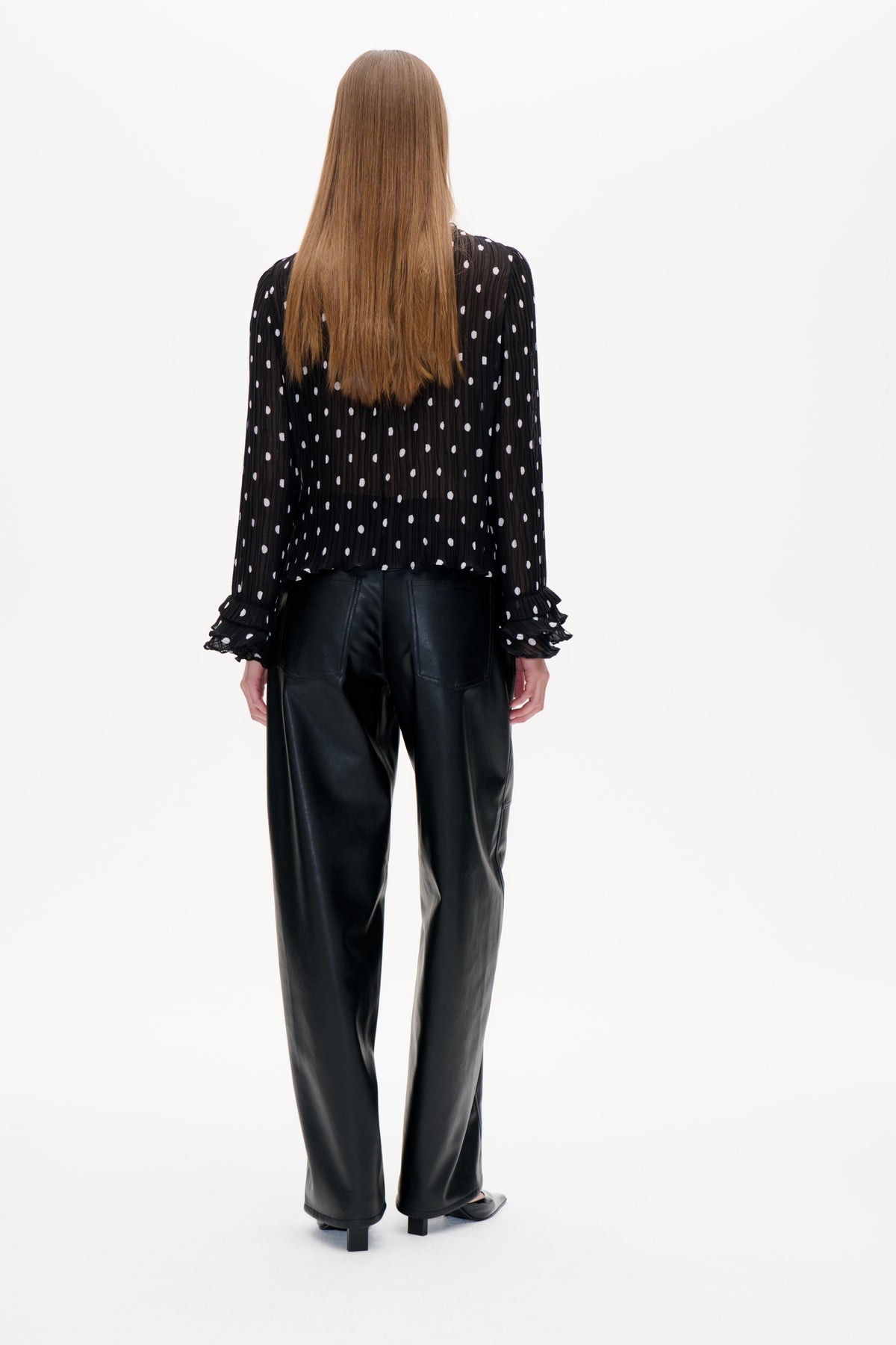 Achteraanzicht van een zwarte, doorschijnende blouse met fijne plooien, witte stippen en gerimpelde manchetten, gecombineerd met een zwarte broek.