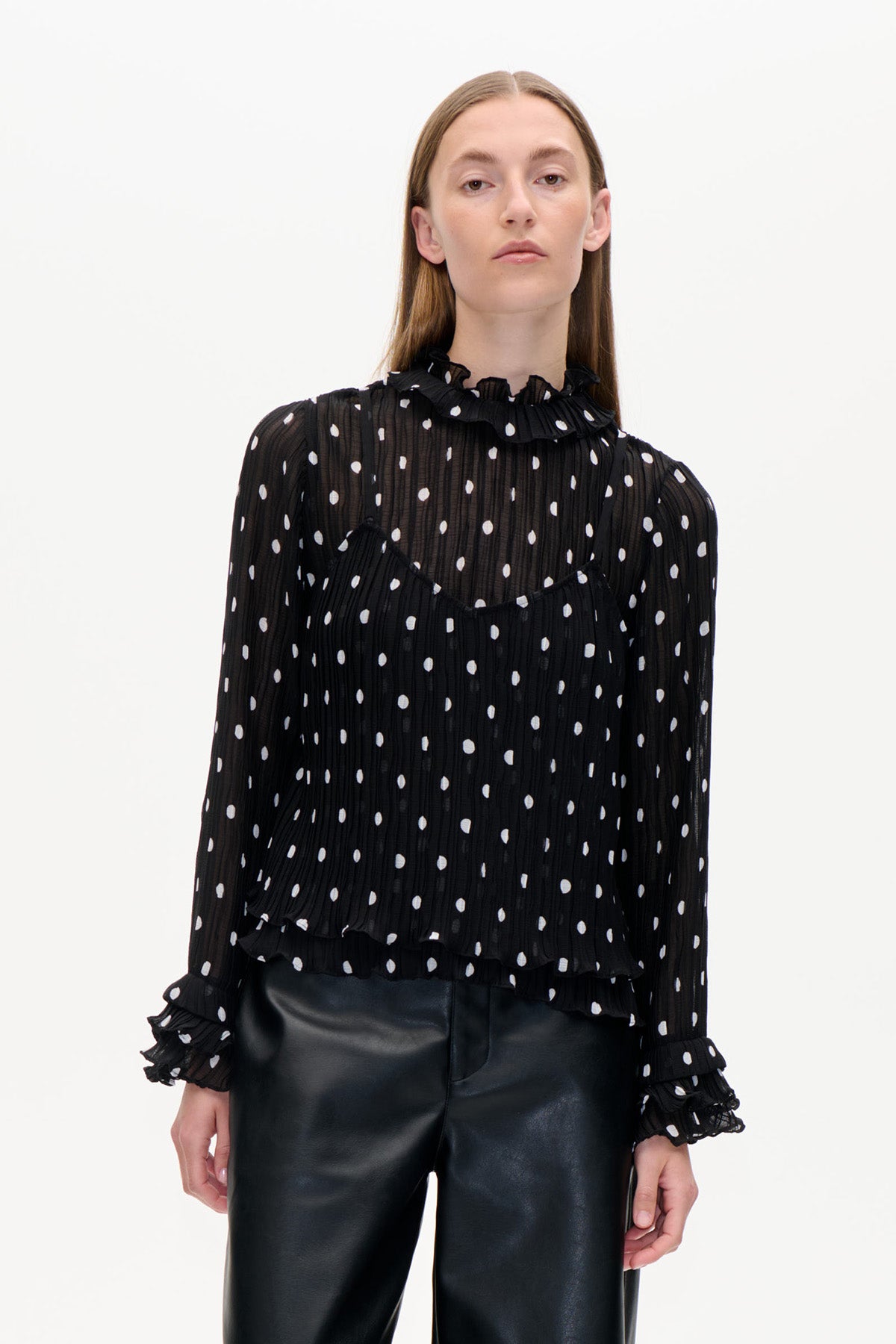 Vooraanzicht van een zwarte, doorschijnende, gestructureerde blouse met witte stippen, ruches bij de hoge kraag en de manchetten, gecombineerd met een zwarte broek.