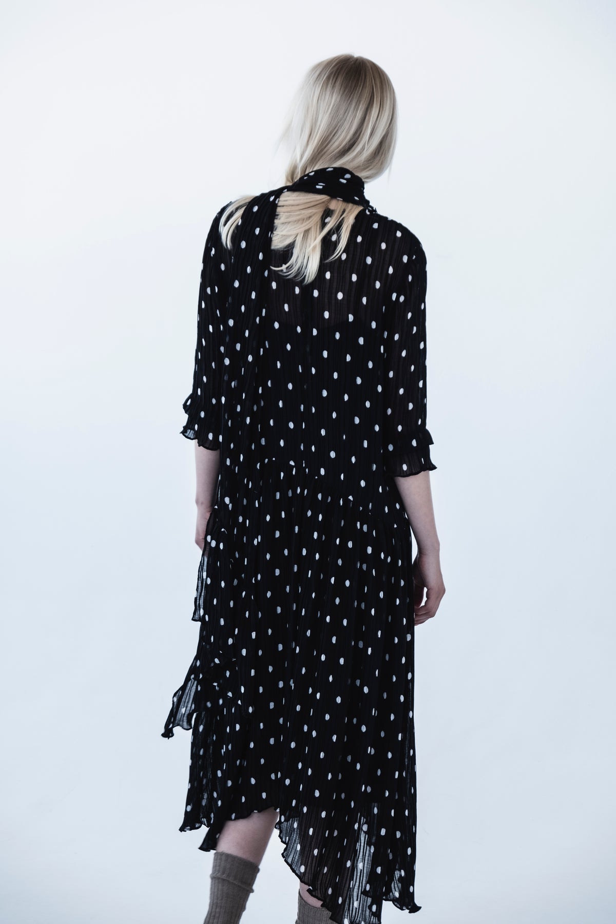 Rear view of a model wearing a black pleated dress with irregular white dots, flutter sleeves and a layered sheer hem against a pale backdrop.