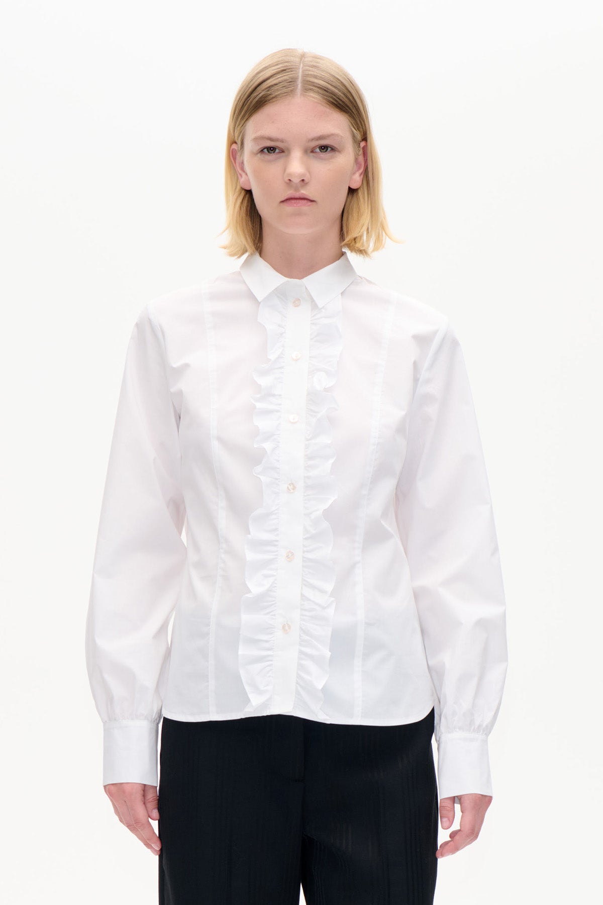 Front view of a white long-sleeved button-up shirt with a pointed collar and ruffled trim along the placket, worn with black trousers.
