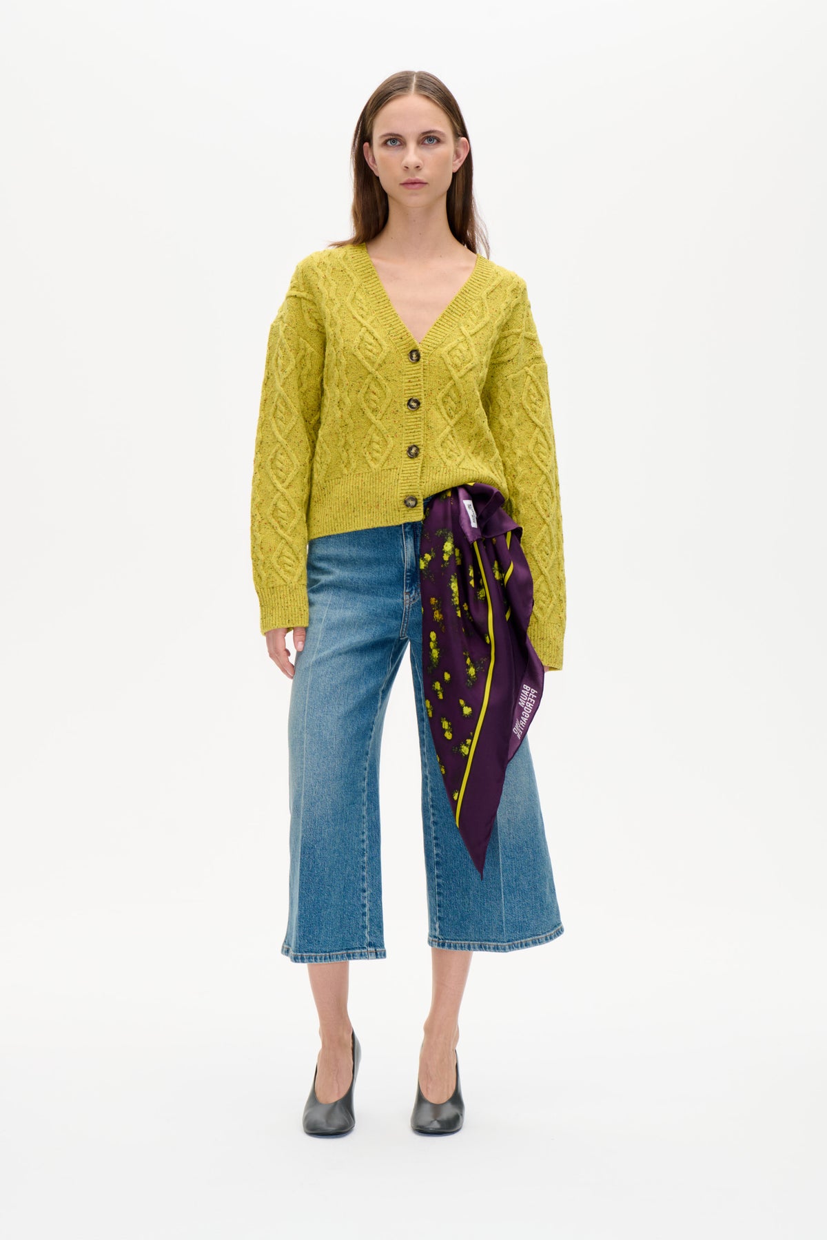 Vorderansicht eines Models, das einen gelbgrünen Zopfstrickcardigan mit Knöpfen, weit geschnittene blaue Jeans und einen lila Blumenschal trägt.