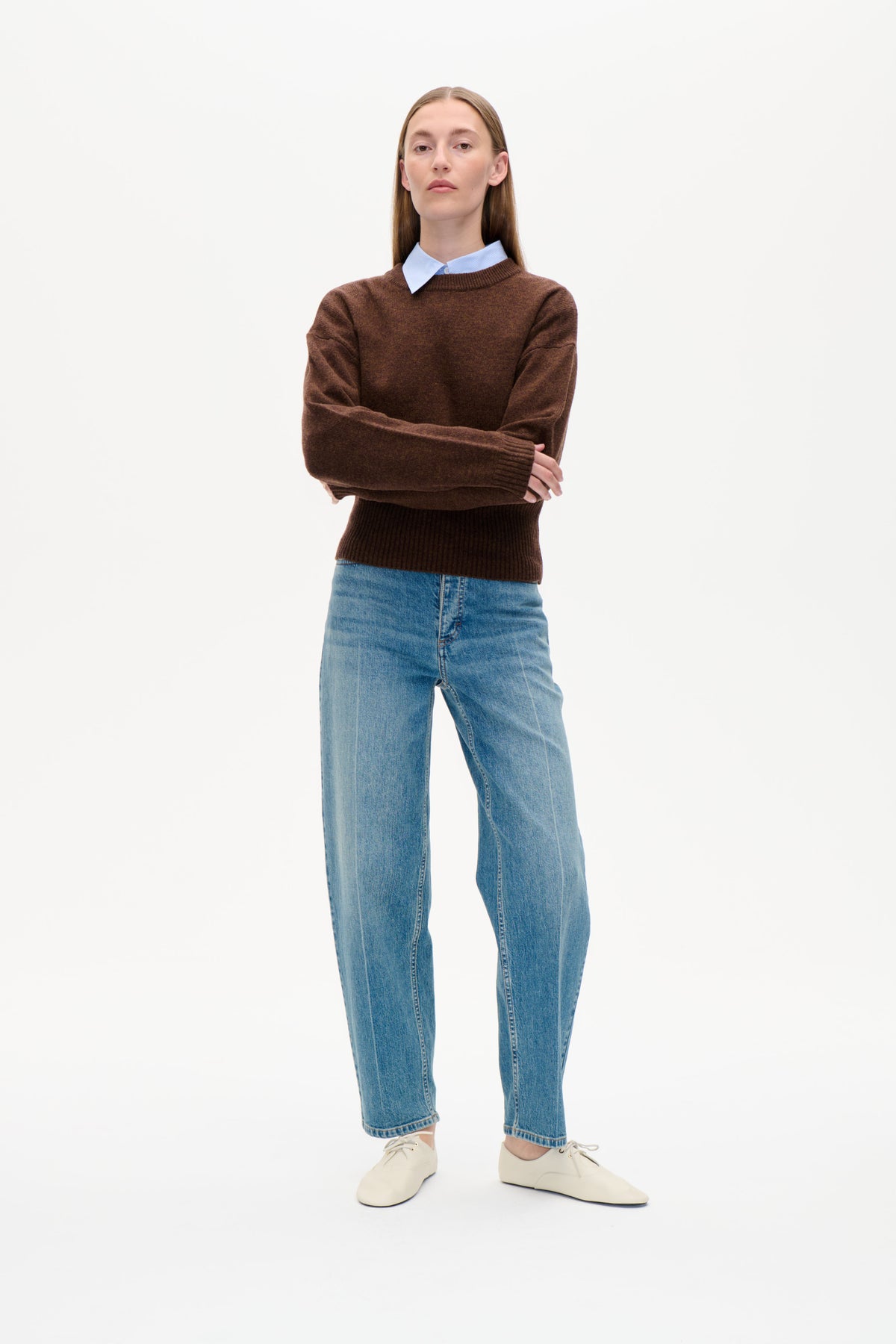 Front view of a model wearing a brown crew-neck sweater over a pale blue striped collared shirt, with blue jeans and cream lace-up shoes.