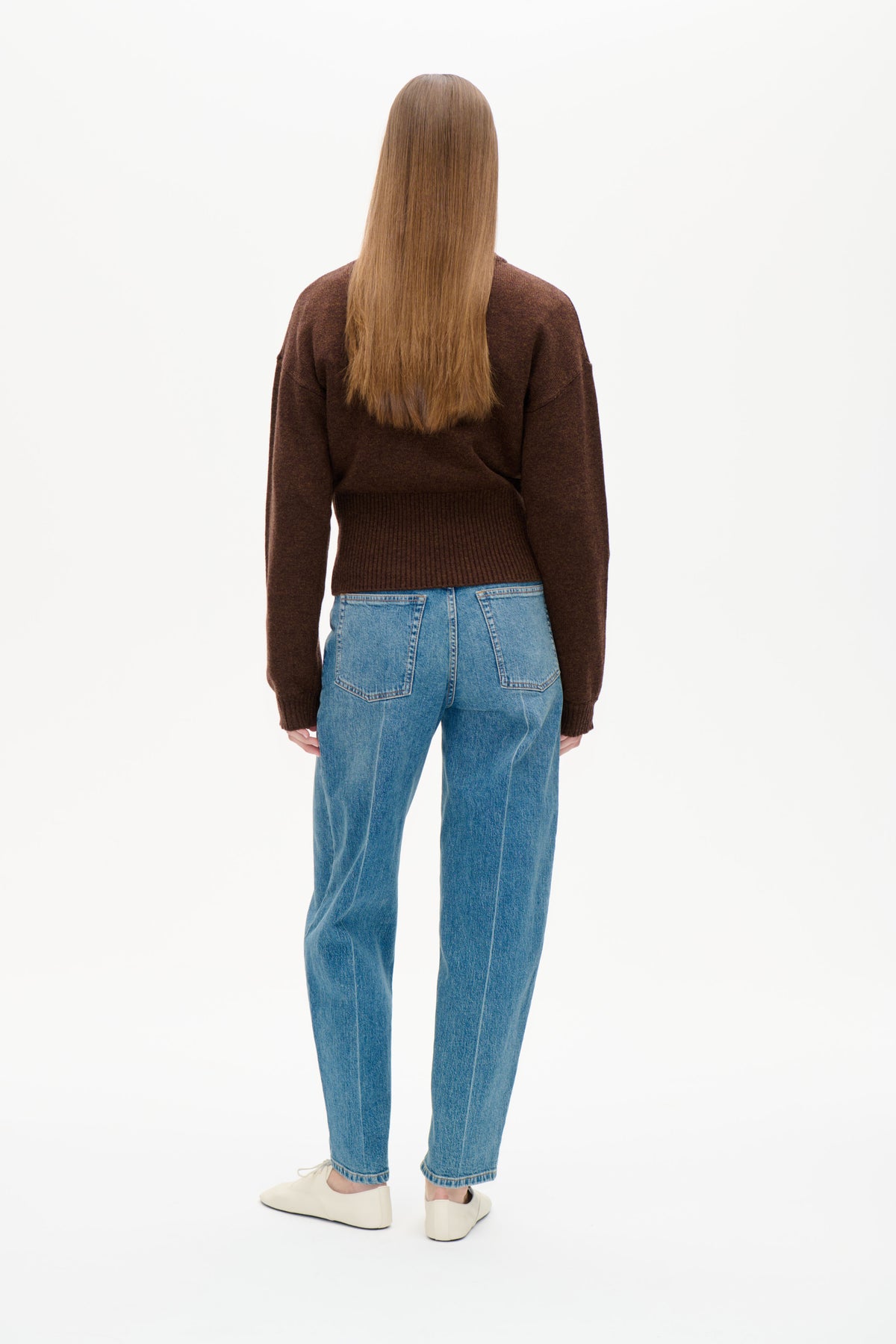 Back view of a model wearing a dark brown long-sleeved knit sweater with a ribbed hem and blue jeans.