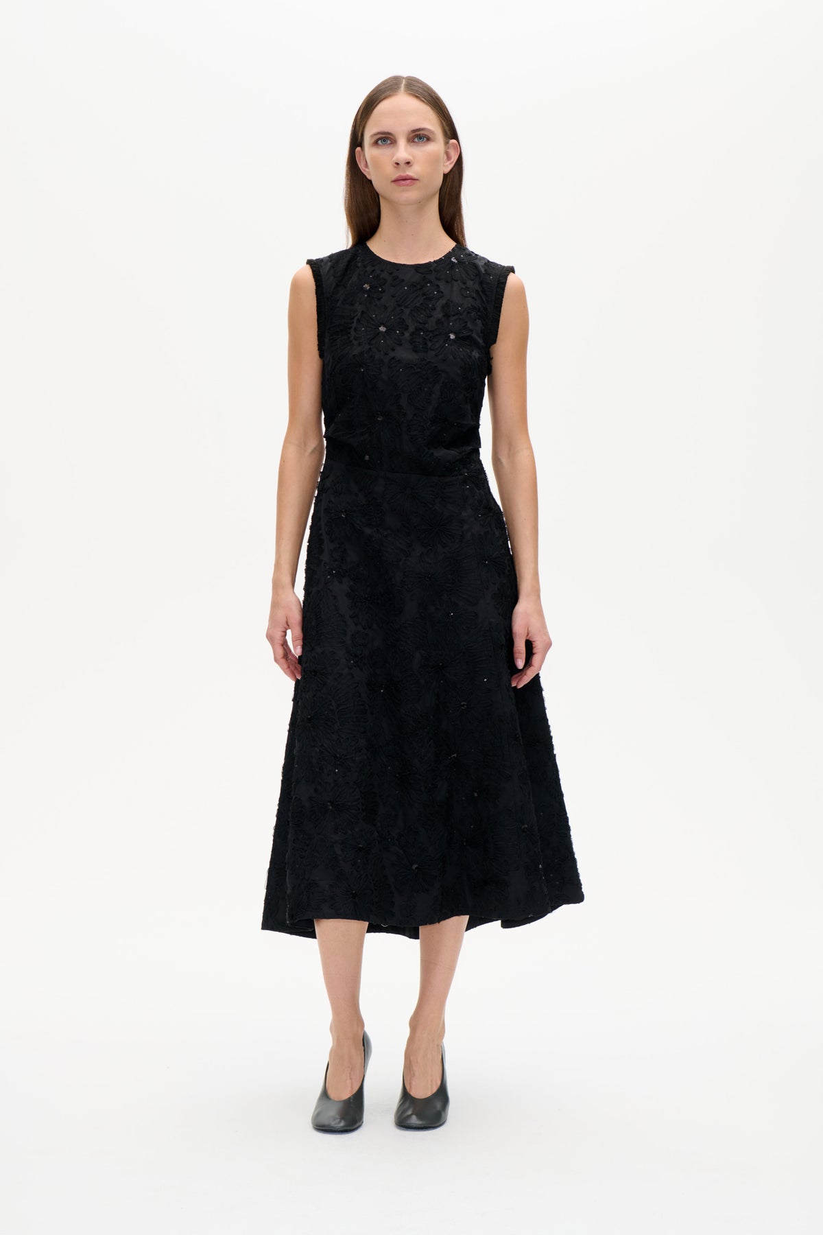 Front view of a model wearing a black sleeveless dress with raised floral details and scattered sequins, fitted waist and flared skirt.