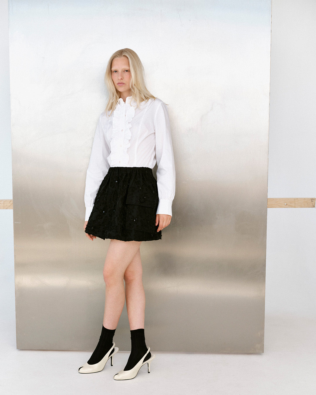 Campaign portrait of a model wearing a white ruffled blouse, black textured mini skirt, black socks and cream high heels.