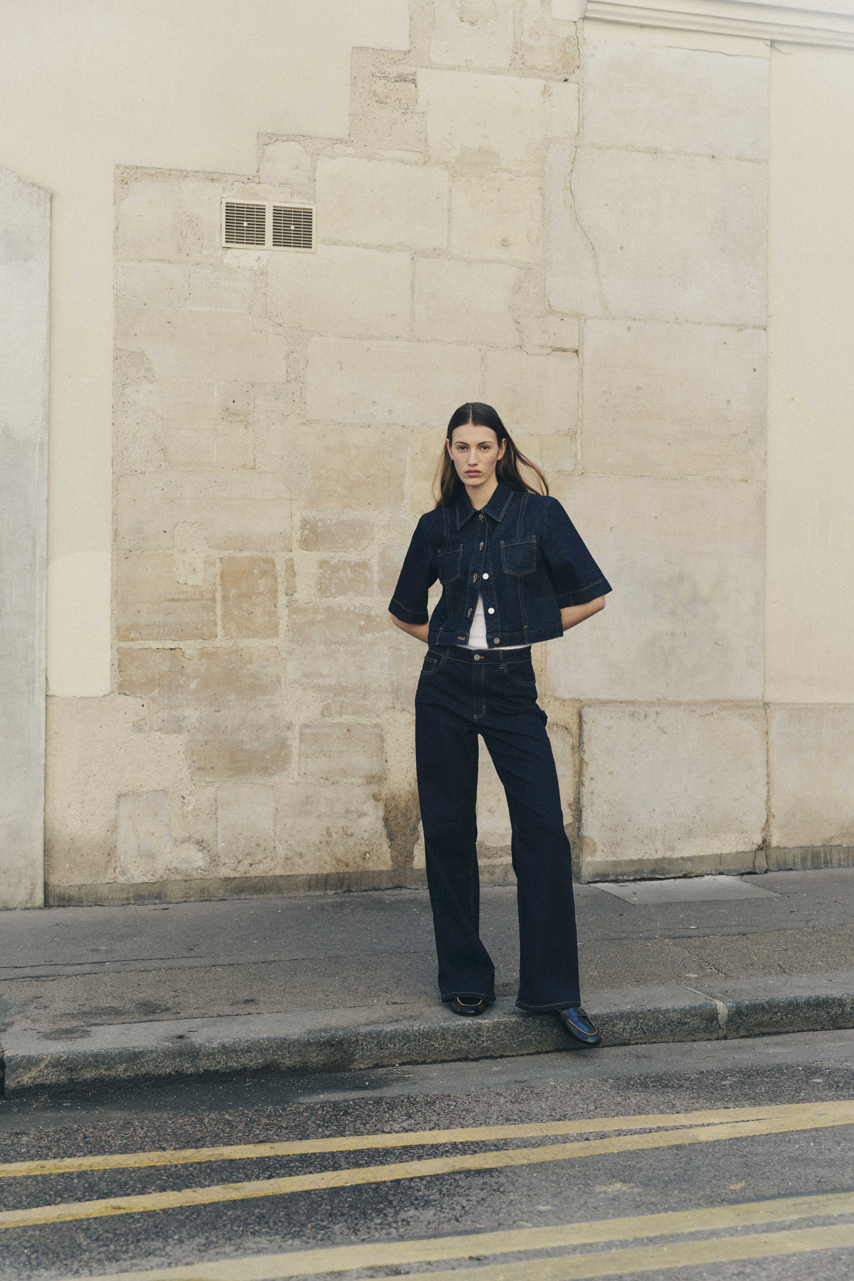 Model wearing a dark indigo denim jacket with contrast stitching and matching wide-leg jeans, standing against a pale stone wall.