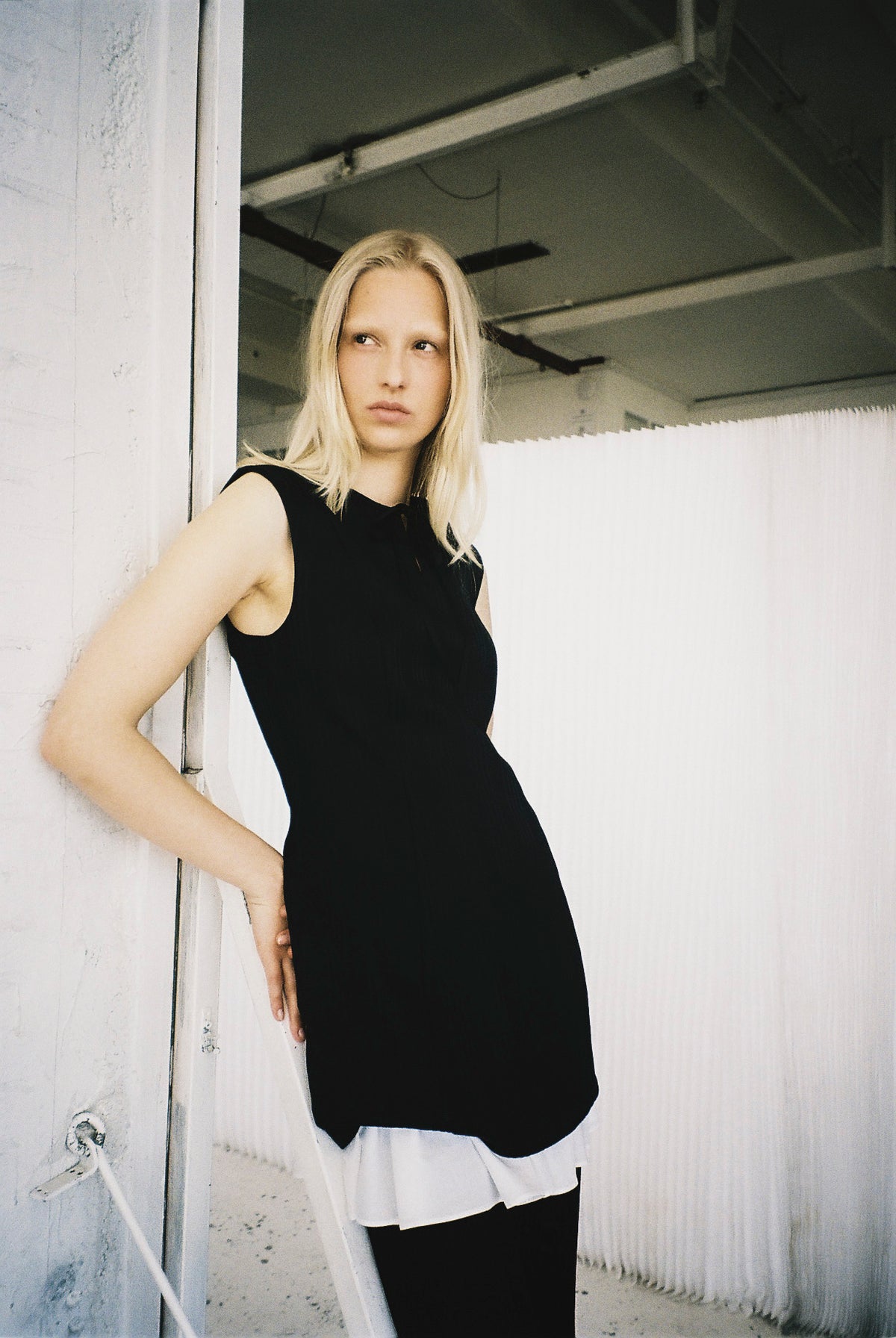 Model wearing a black sleeveless fitted tunic over a white shirt with a ruffled hem, standing beside a white wall.