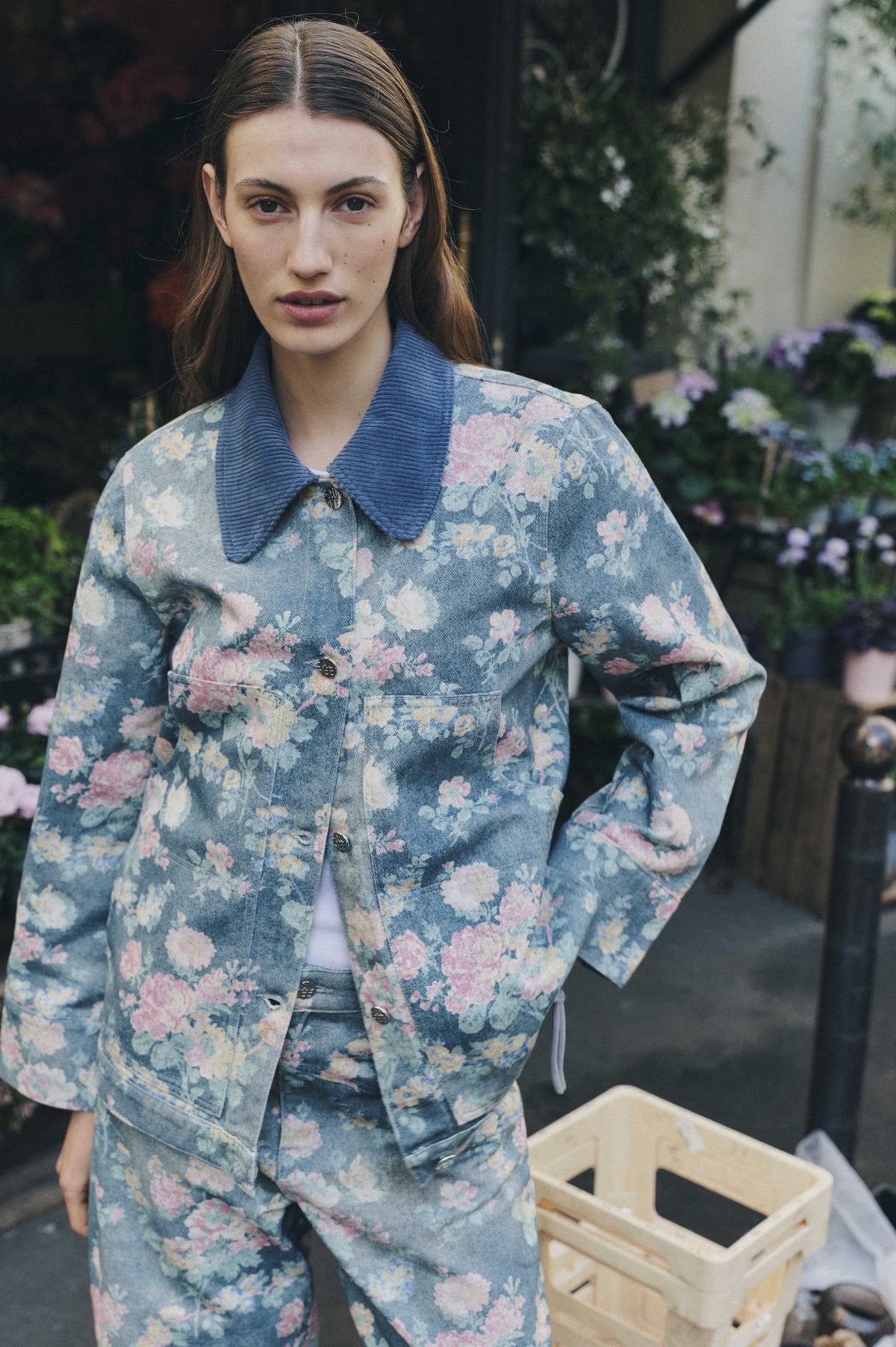 Campagneportret van een model met een blauwe denimjas en bijpassende broek, voorzien van een bloemenprint in roze, wit en geel en een geribde blauwe kraag.
