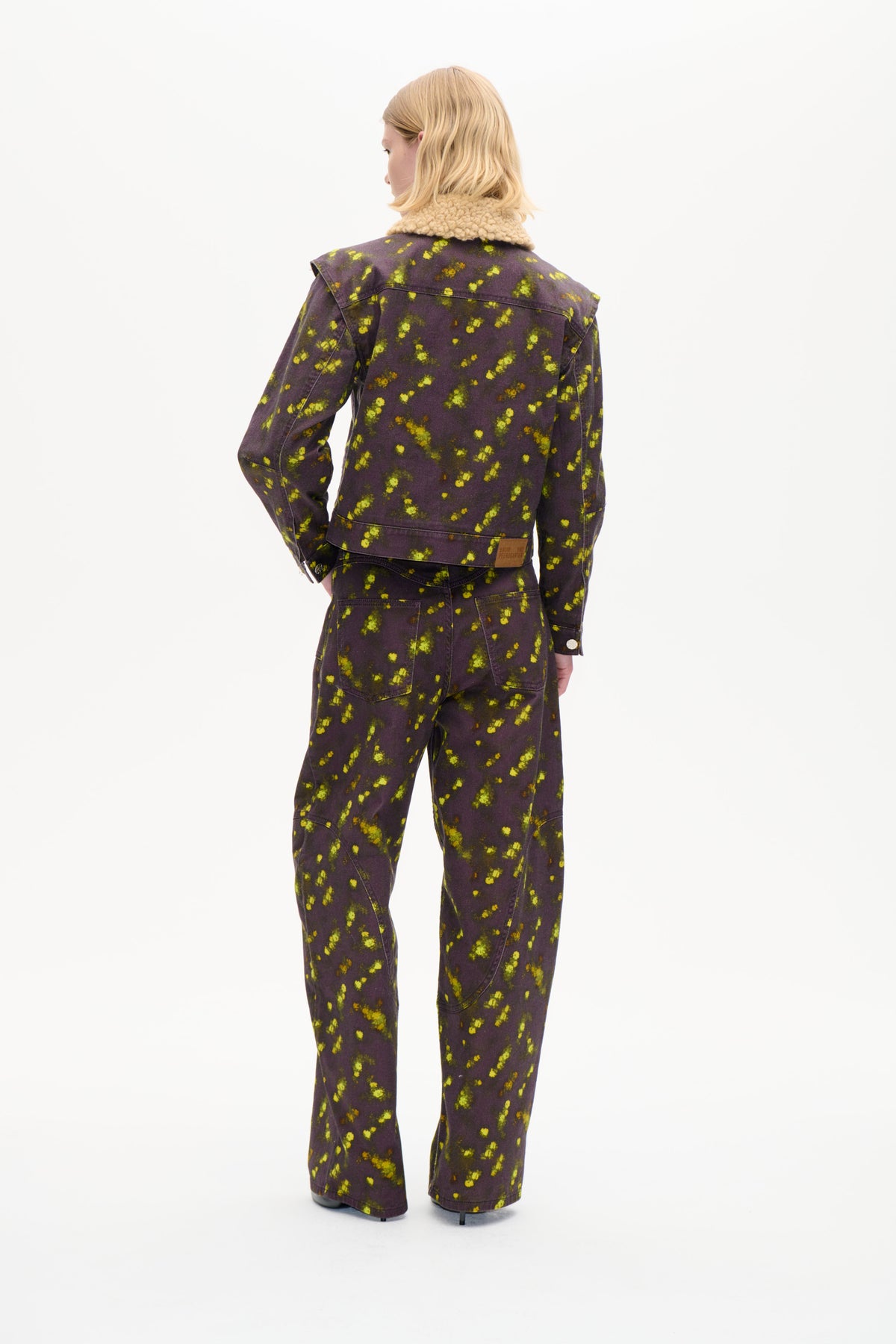 Rückansicht eines Models in einer dunkelpflaumenfarbenen Jacke mit dickem cremefarbenem Kragen und einer Hose mit verstreutem limonengelben Blumenprint.