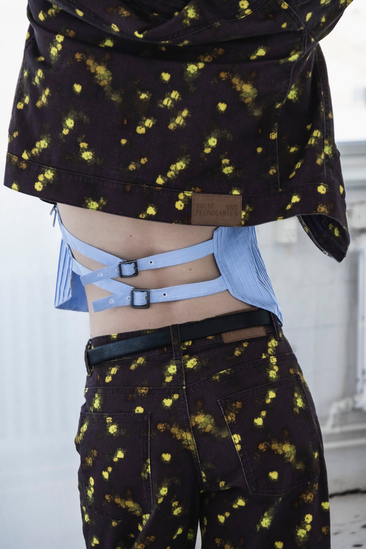 Nahaufnahme des Kampagnen-Stylings: dunkle denim-Jacke und passende Hose mit gelb-grünem Blumenprint, darüber eine blau-gestreifte Cut-out-Bluse mit Schnallenriemen.